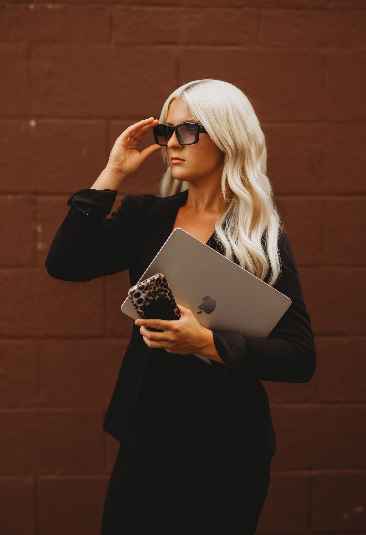 Blonde woman in black blazer holding MacBook and leopard print clutch, wearing sunglasses against brick wall.
