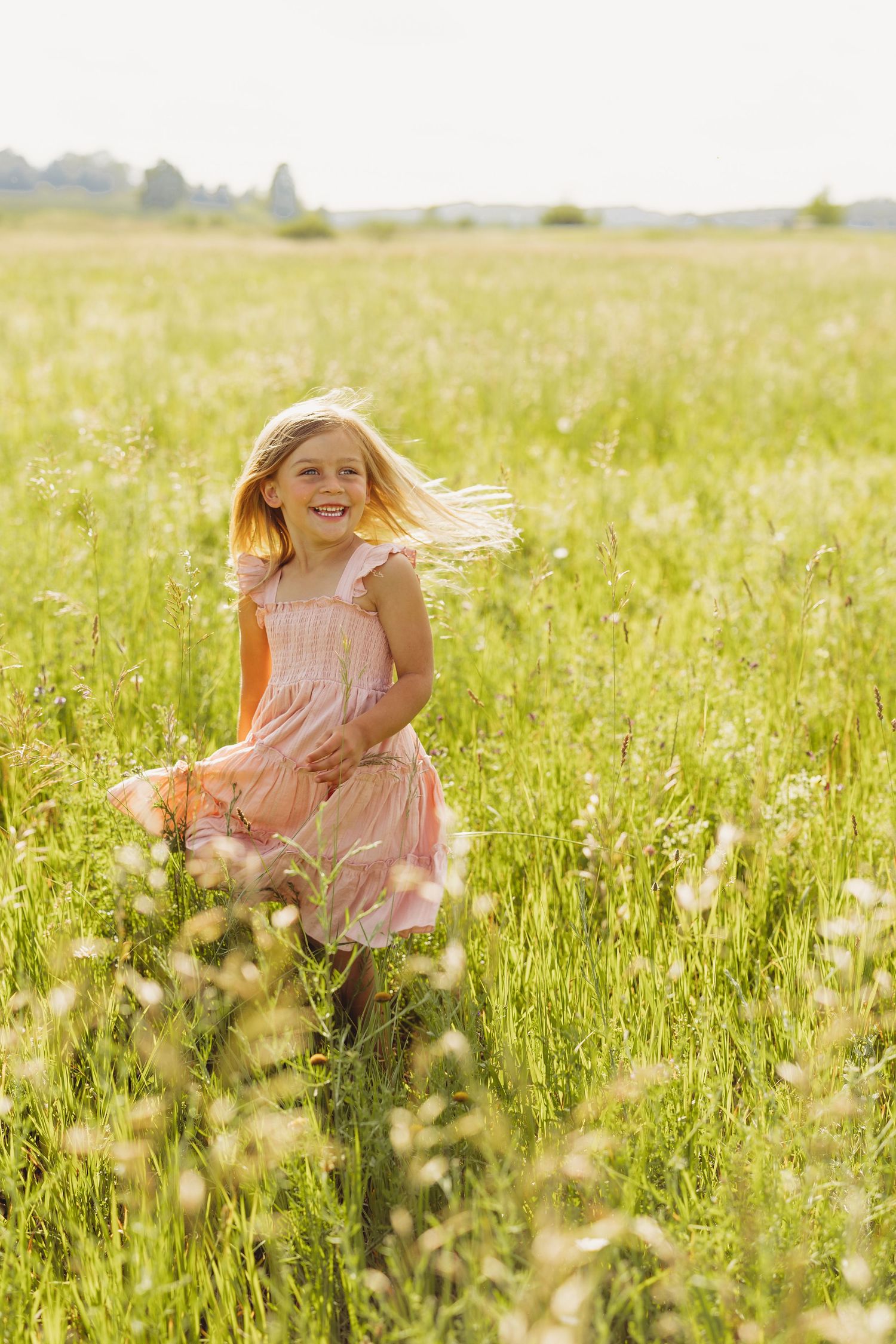A joyful blonde girl in a pink dress runs through a sunny green meadow with wildflowers on a bright summer day.