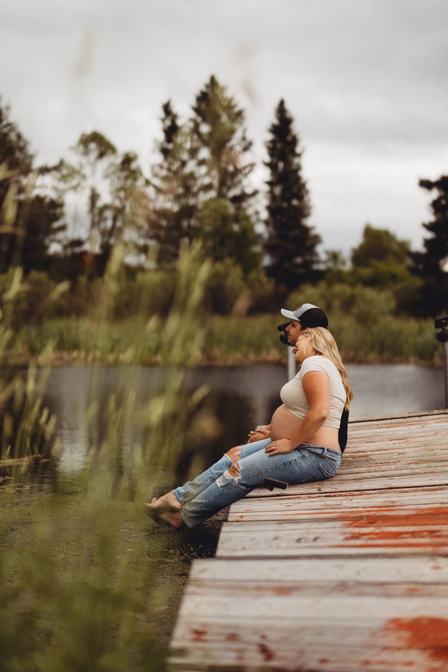 Expecting couple sitting on a rustic dock by a serene lake, embracing and cradling baby bump.