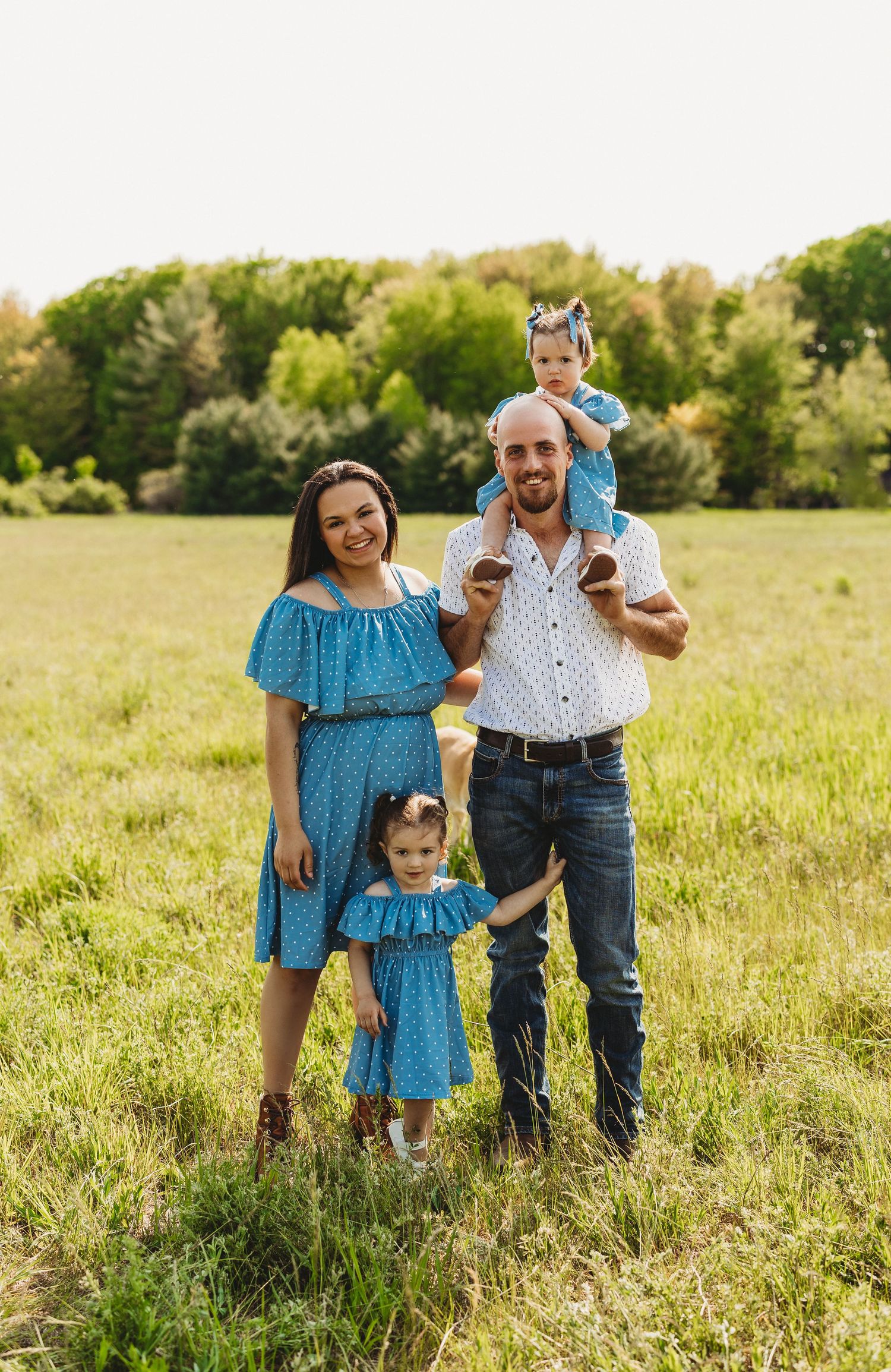 Family wearing coordinating blue and white outfits poses together in a sunny meadow during golden hour.