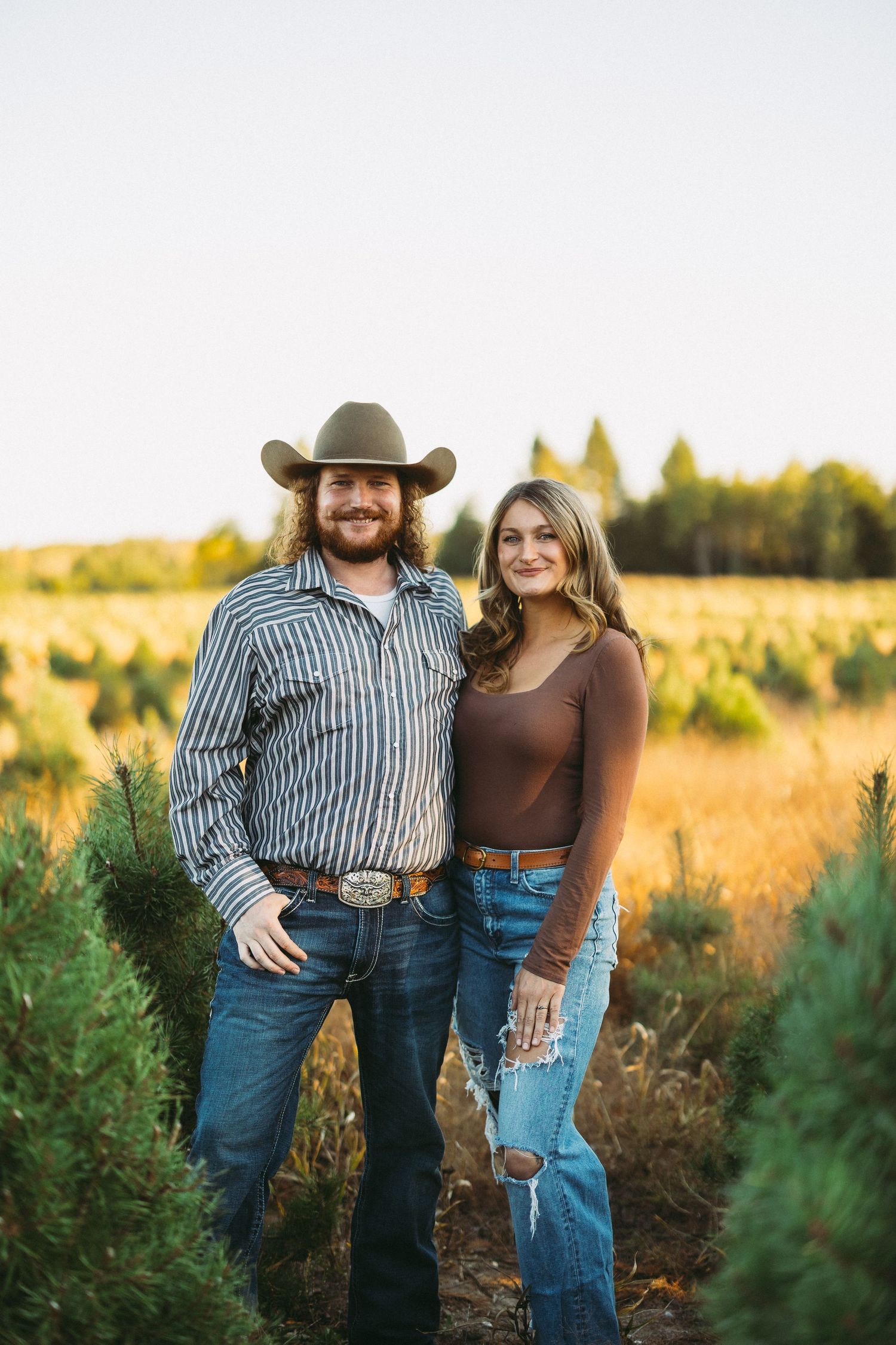 Couple in western wear posing together in field at sunset.
