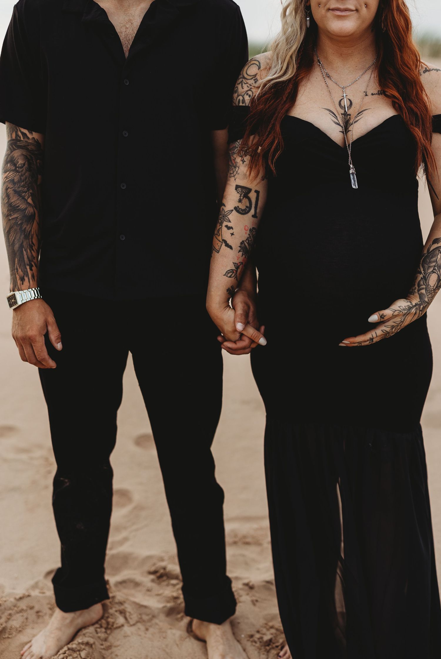 Couple in matching black outfits holding hands on beach showing tattoo sleeves.