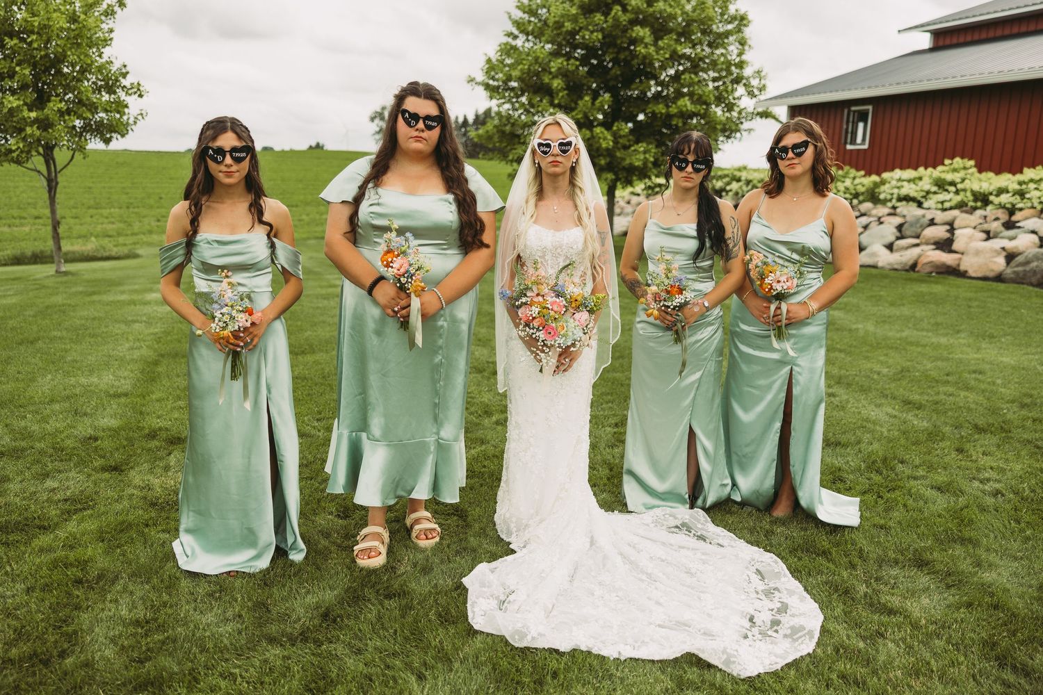 Bridal party in mint green dresses poses together on grassy field near red barn.