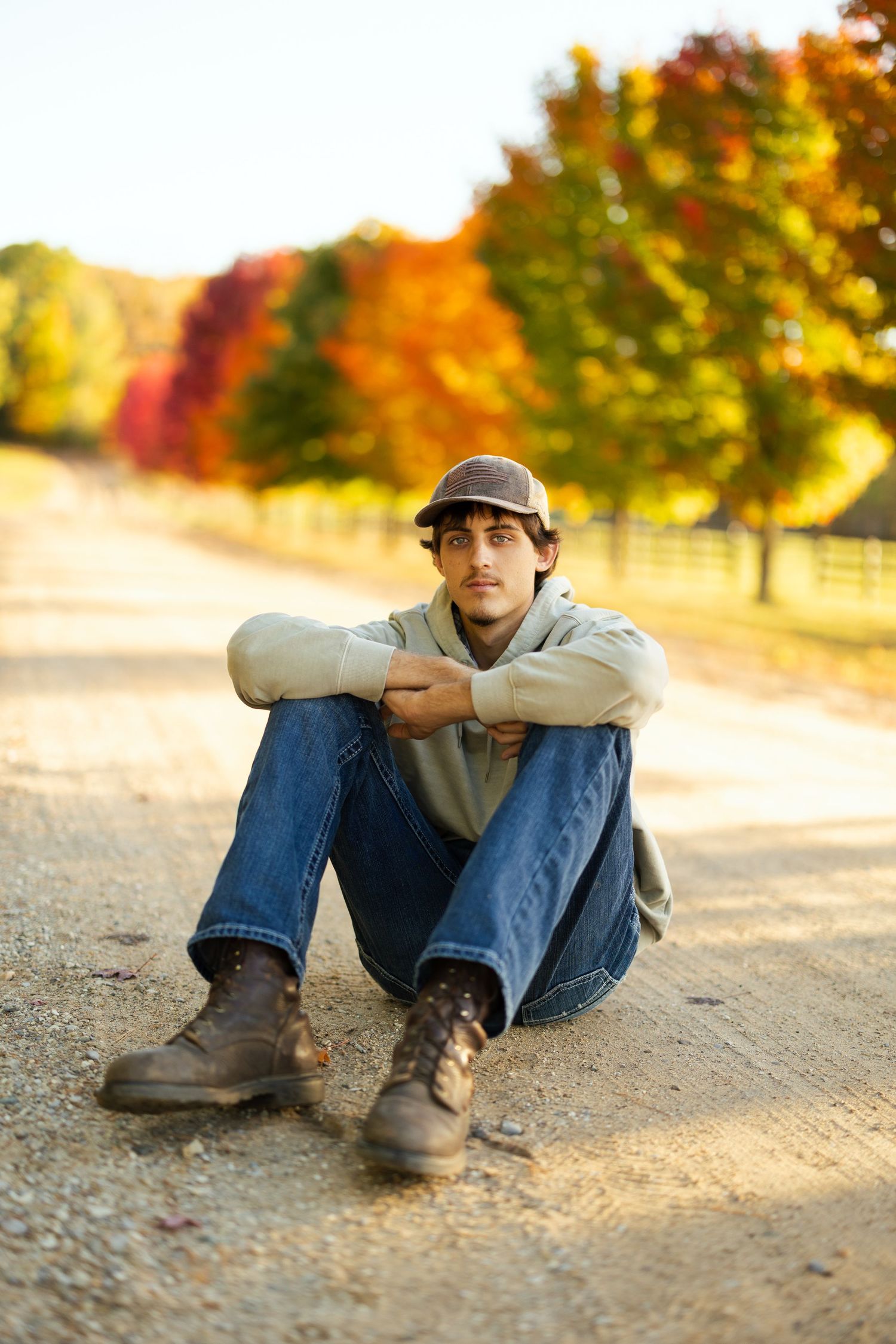 Person sitting on dirt path wearing casual clothing with autumn foliage in background.