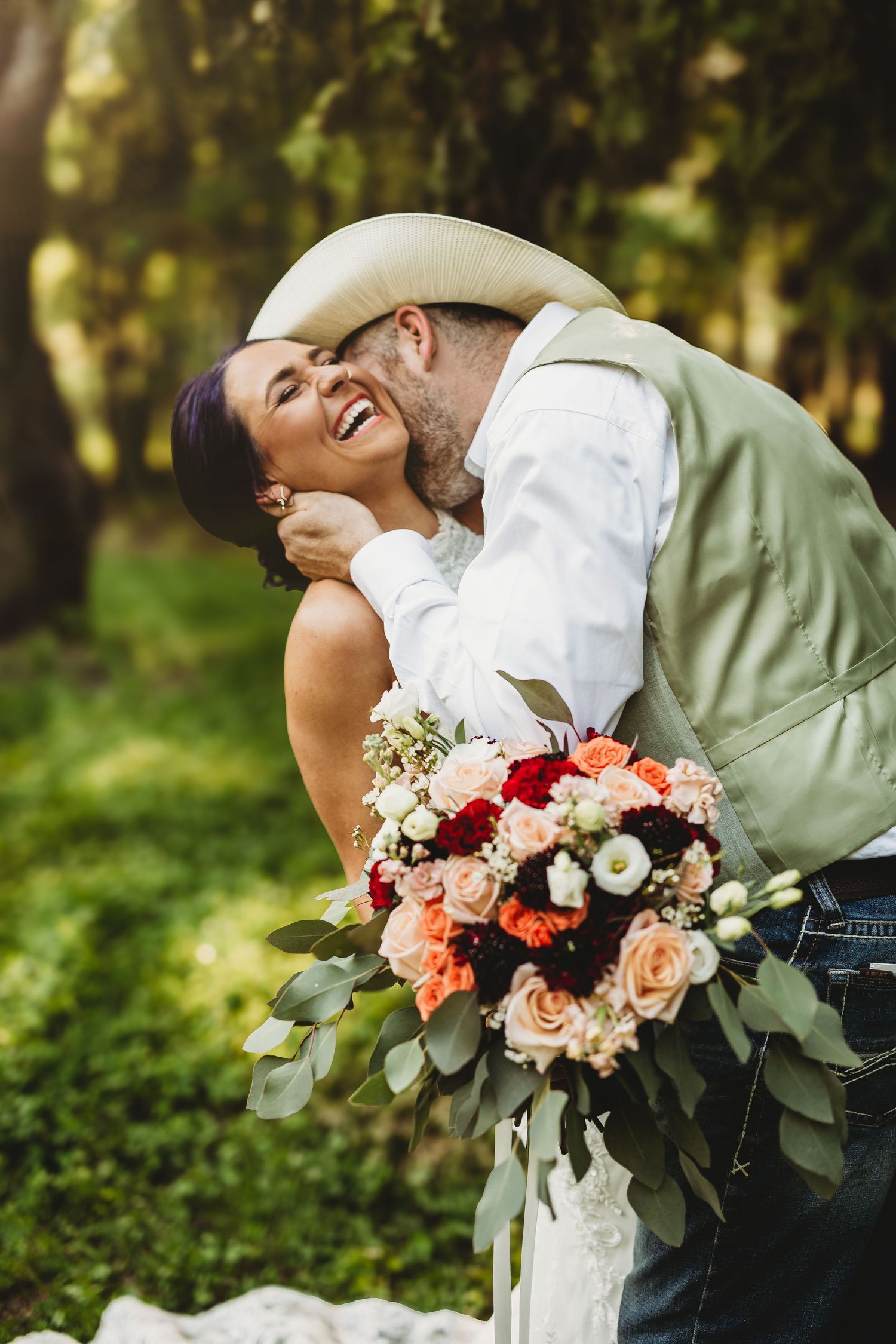 A joyful wedding couple shares a romantic moment outdoors with a beautiful bouquet of roses and greenery.