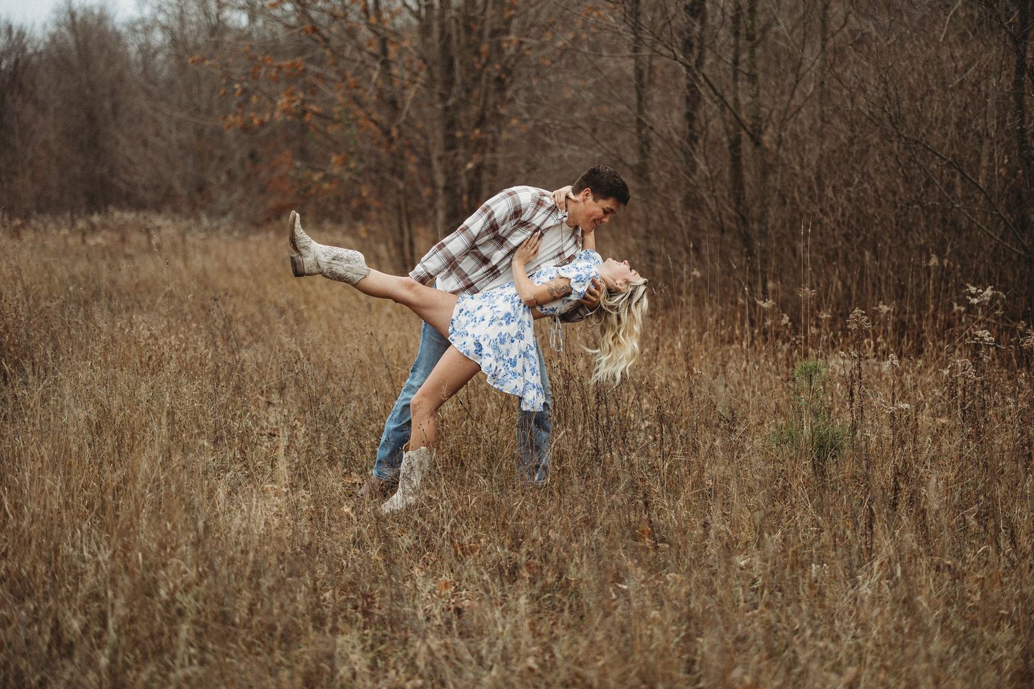A couple in plaid and denim share a playful dance move in a rustic autumn field surrounded by bare trees.