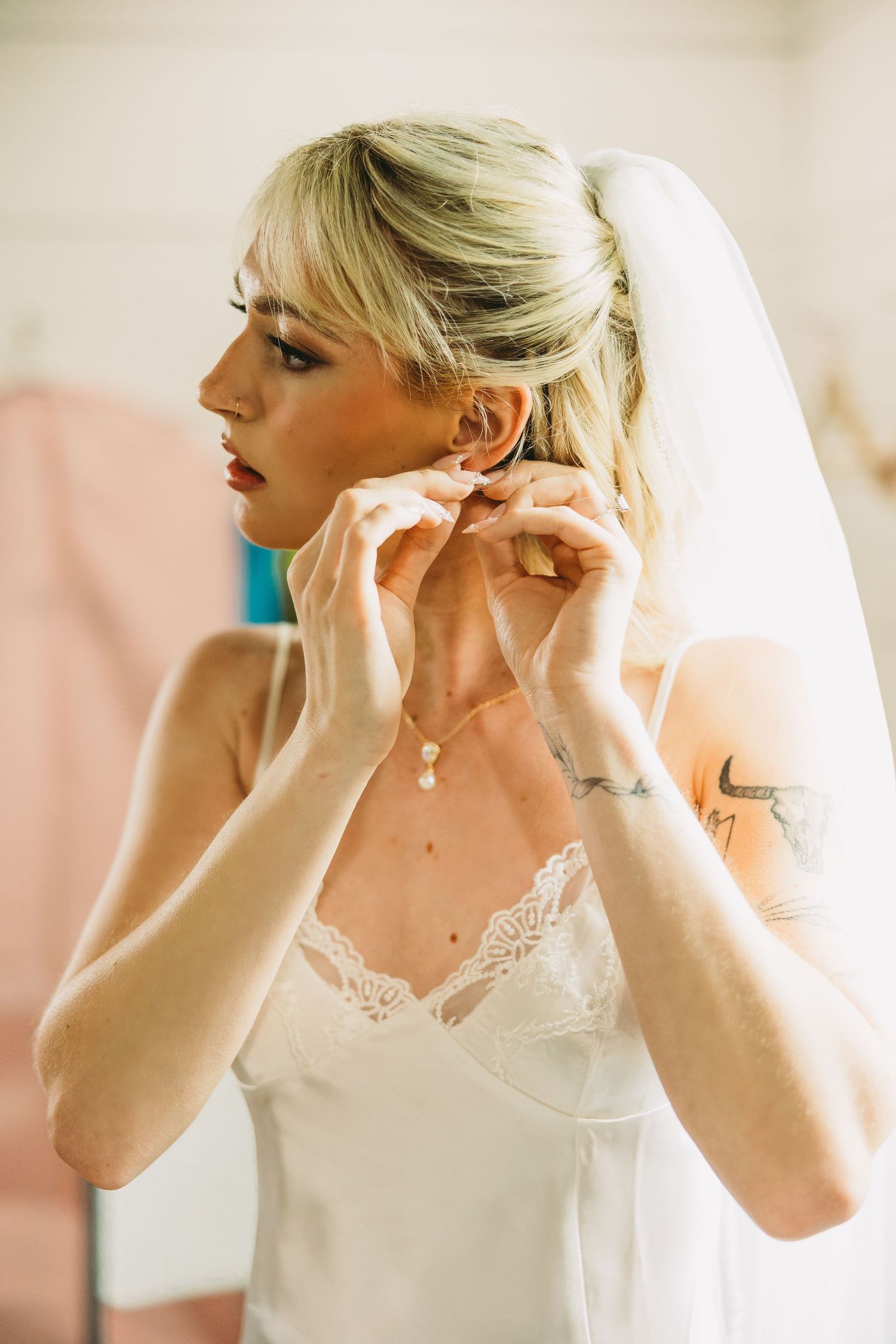 A bride puts on delicate jewelry while getting ready in a soft, natural light setting.