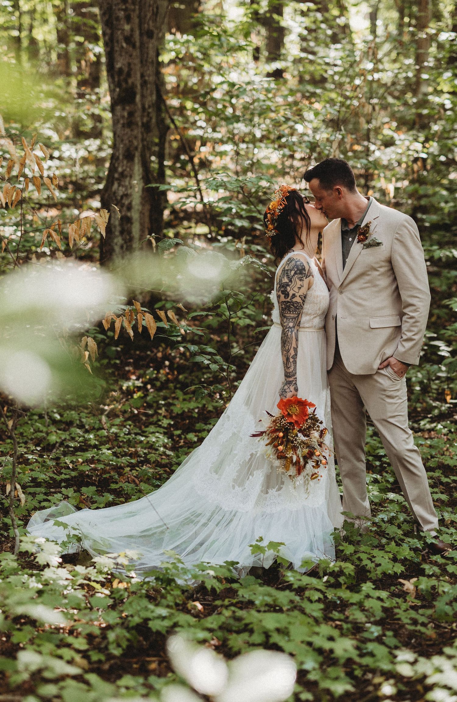 A romantic wedding couple embraces in a lush forest setting surrounded by greenery and white flowers.