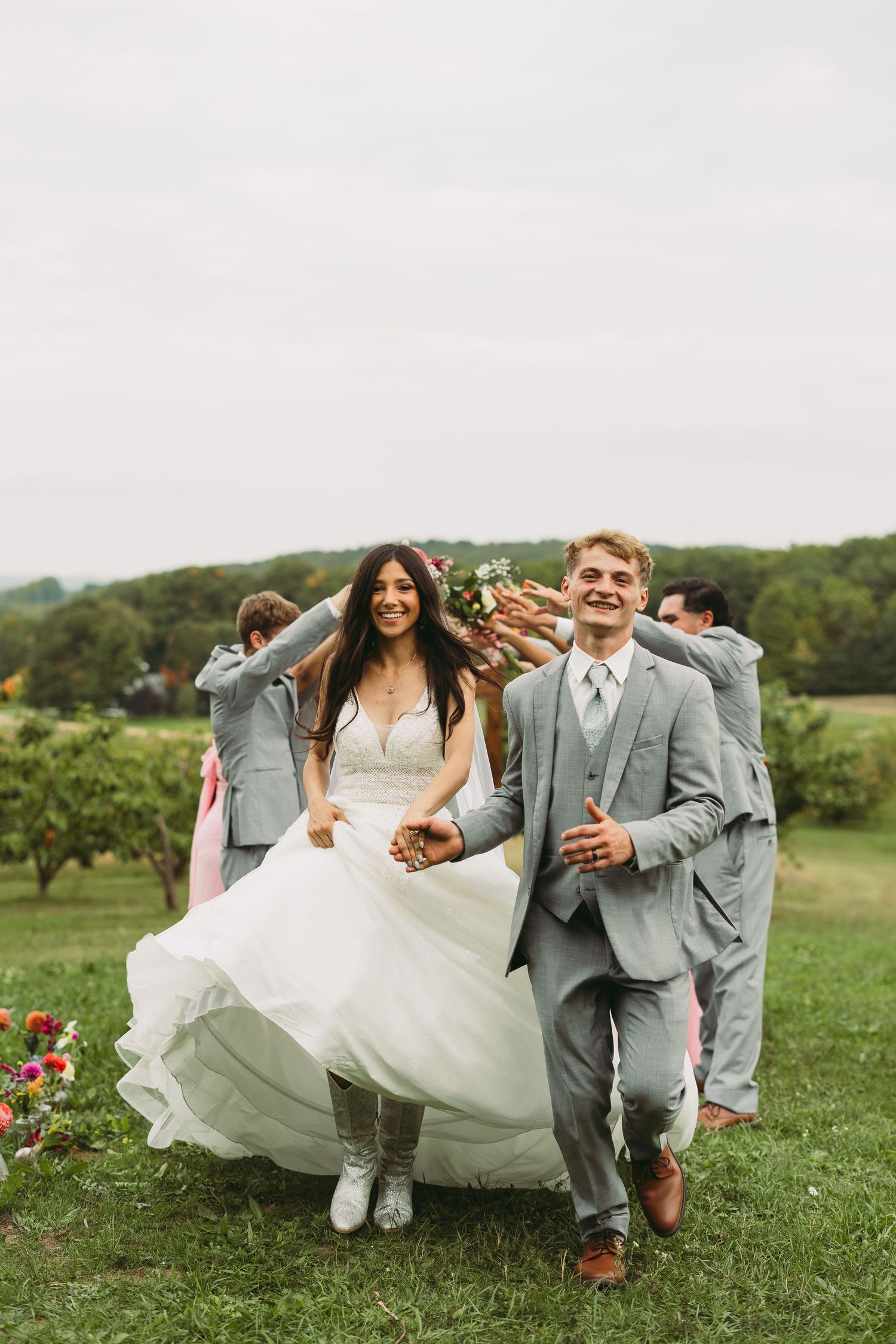 Wedding party members in gray suits and white dress walk playfully through a green field on a sunny day.