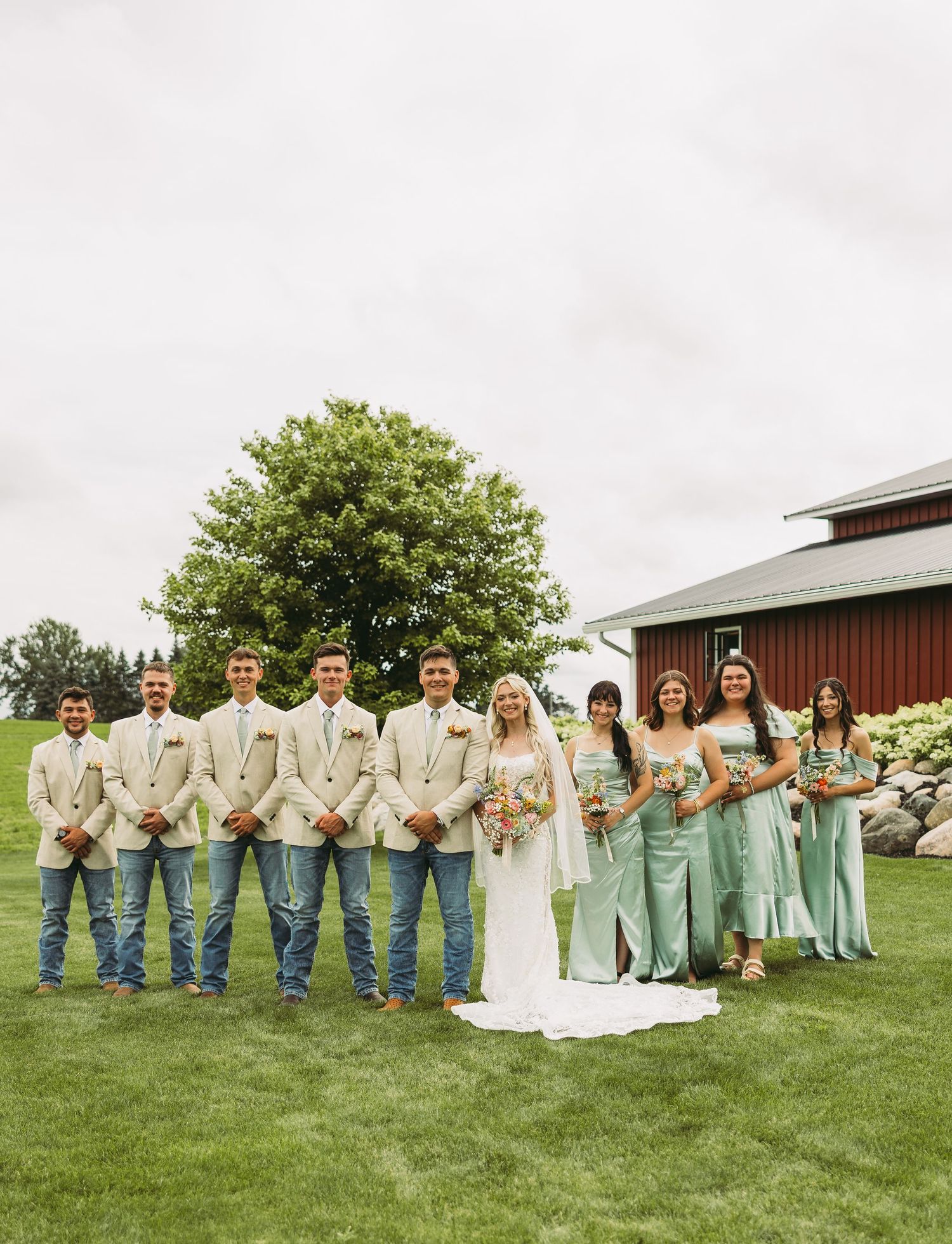 Wedding party in mint green dresses and tan suits poses on grass in front of a red barn.