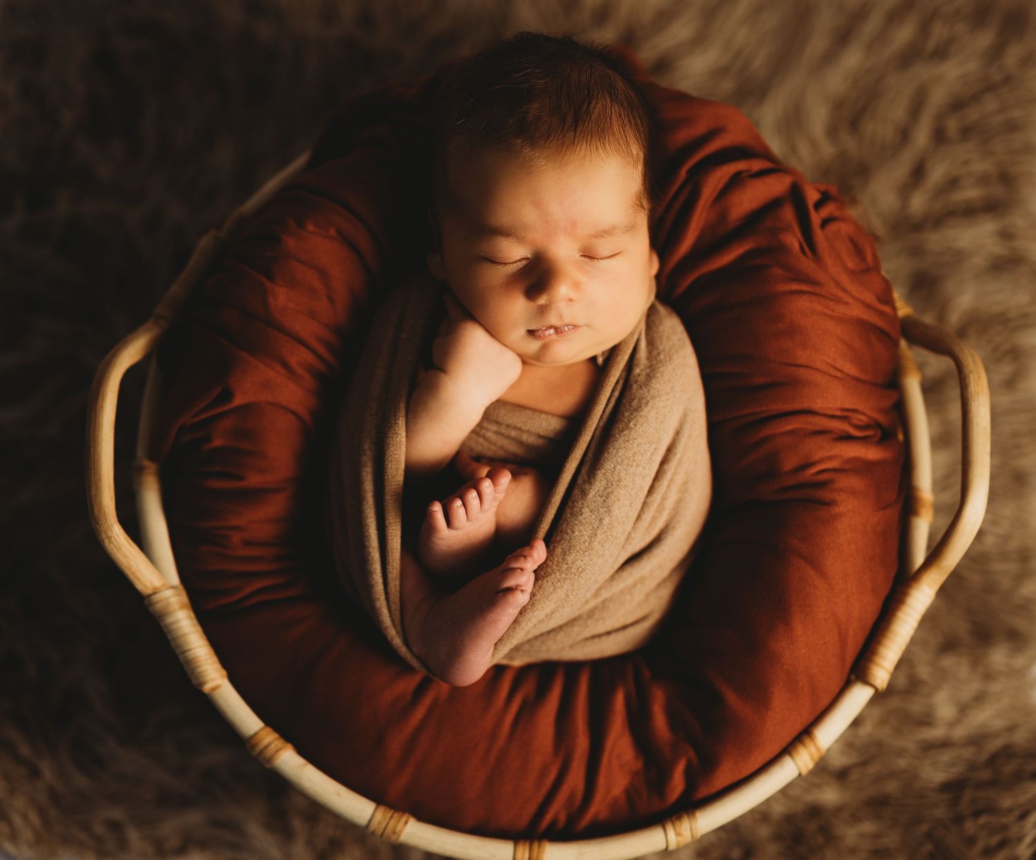 Sleeping newborn wrapped in brown fabric nestled in round basket with dramatic lighting.