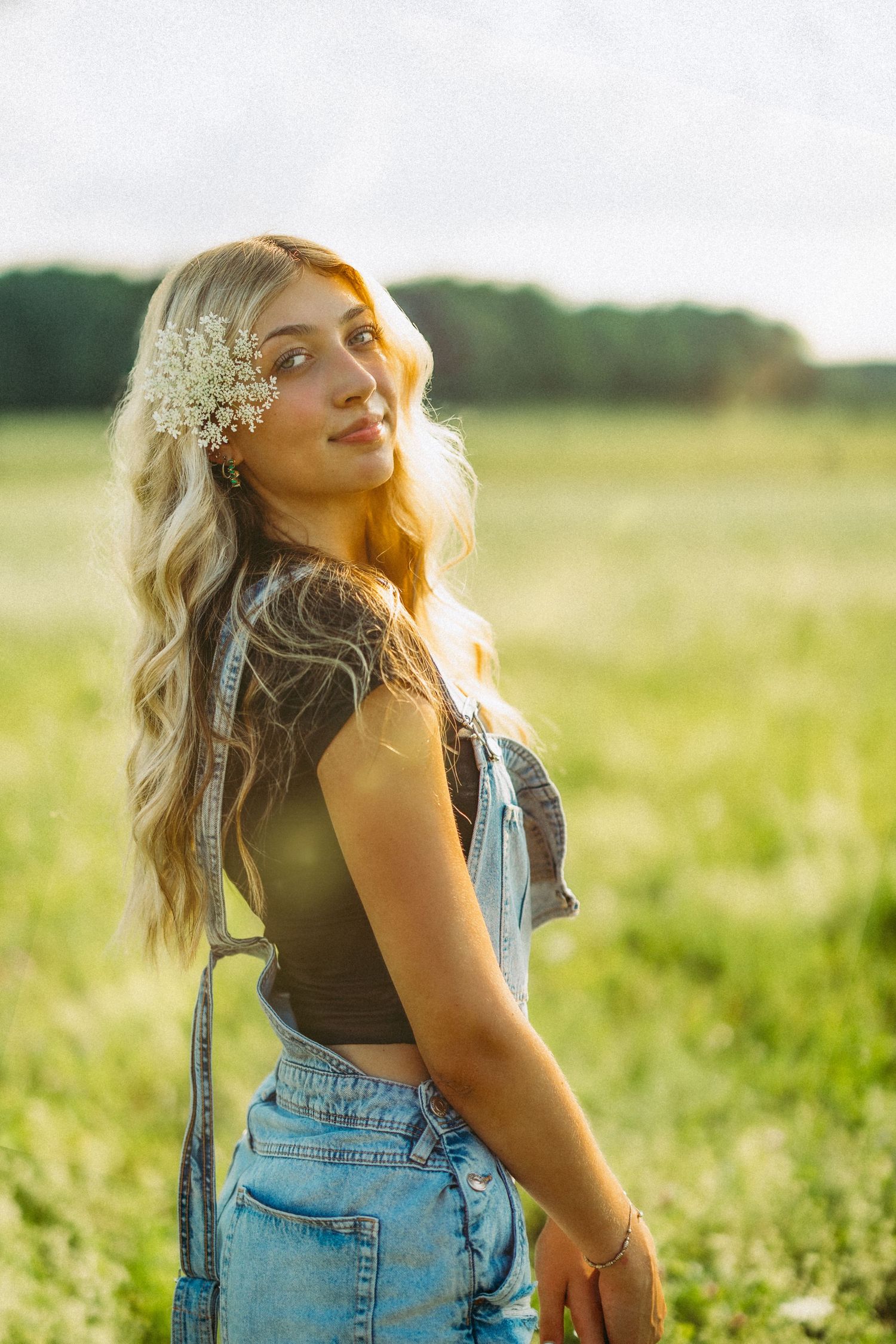 Someone in denim and flowers in their hair poses in a golden-lit meadow at sunset.