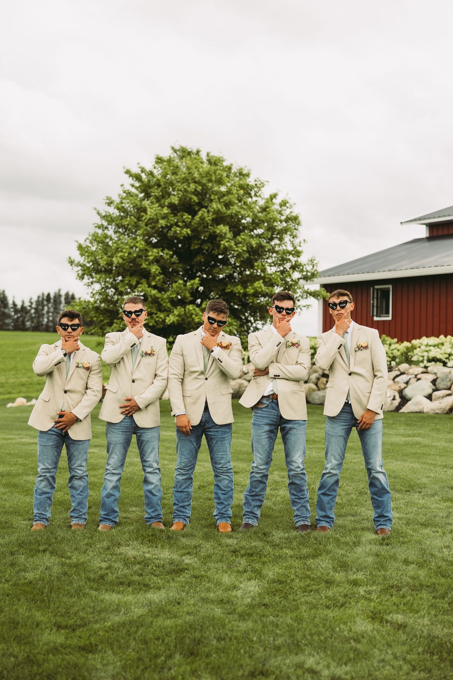 Groomsmen in beige blazers and jeans pose casually on green lawn with red barn in background.