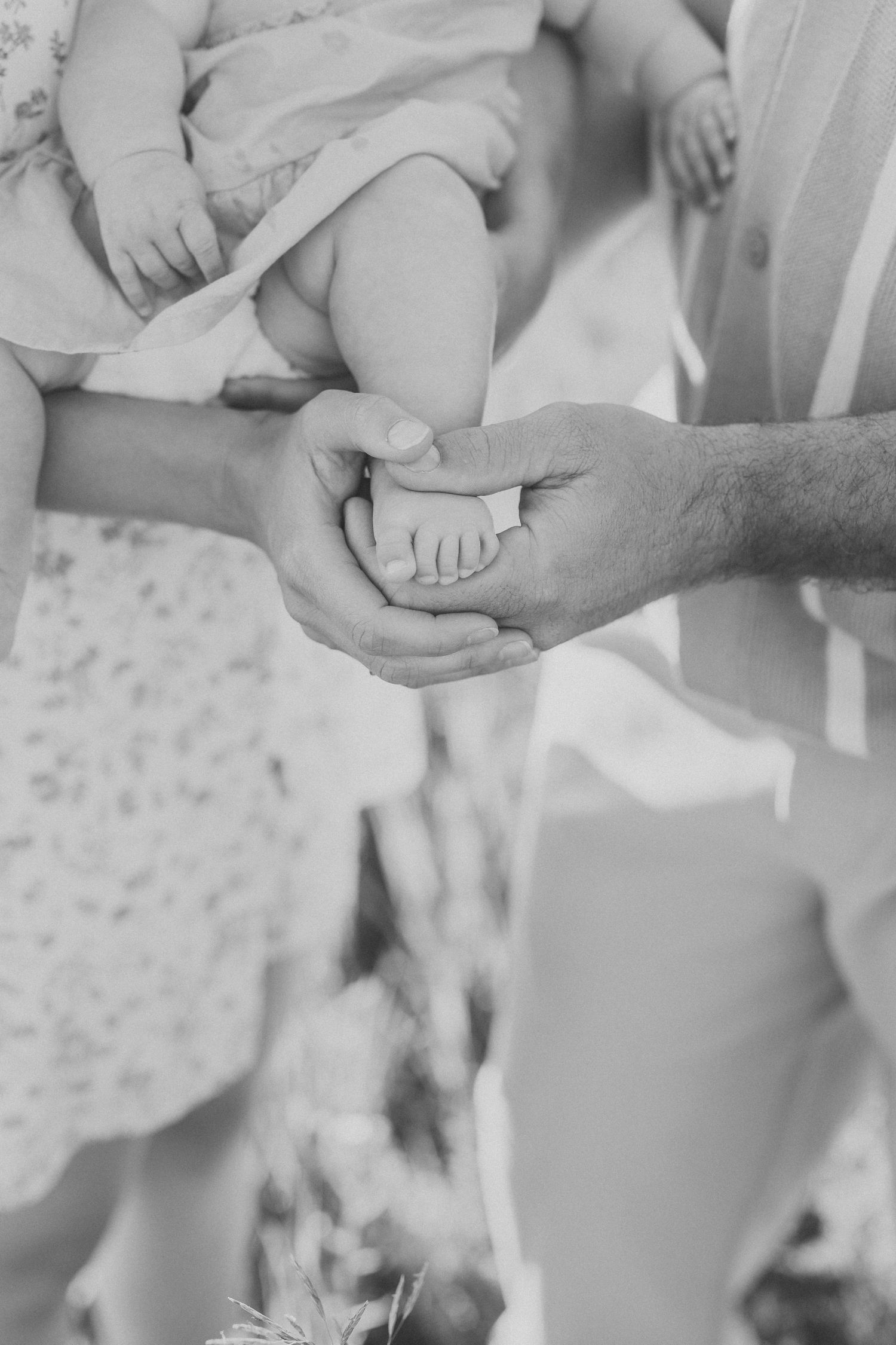 Black and white photo of parents cradling their newborn baby's tiny feet in their hands outdoors.
