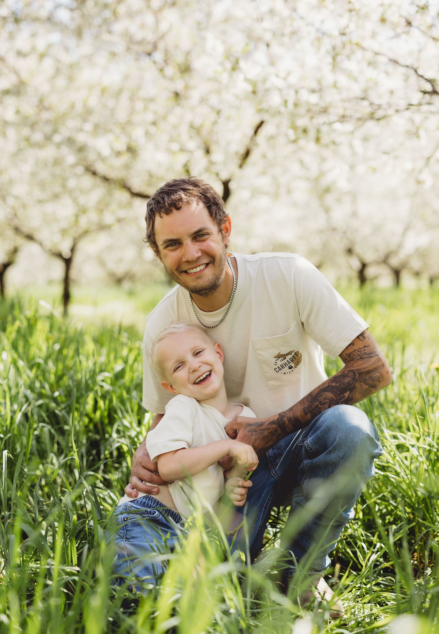 Smiling father crouching with laughing toddler in green grass beneath blooming white cherry trees in spring orchard.