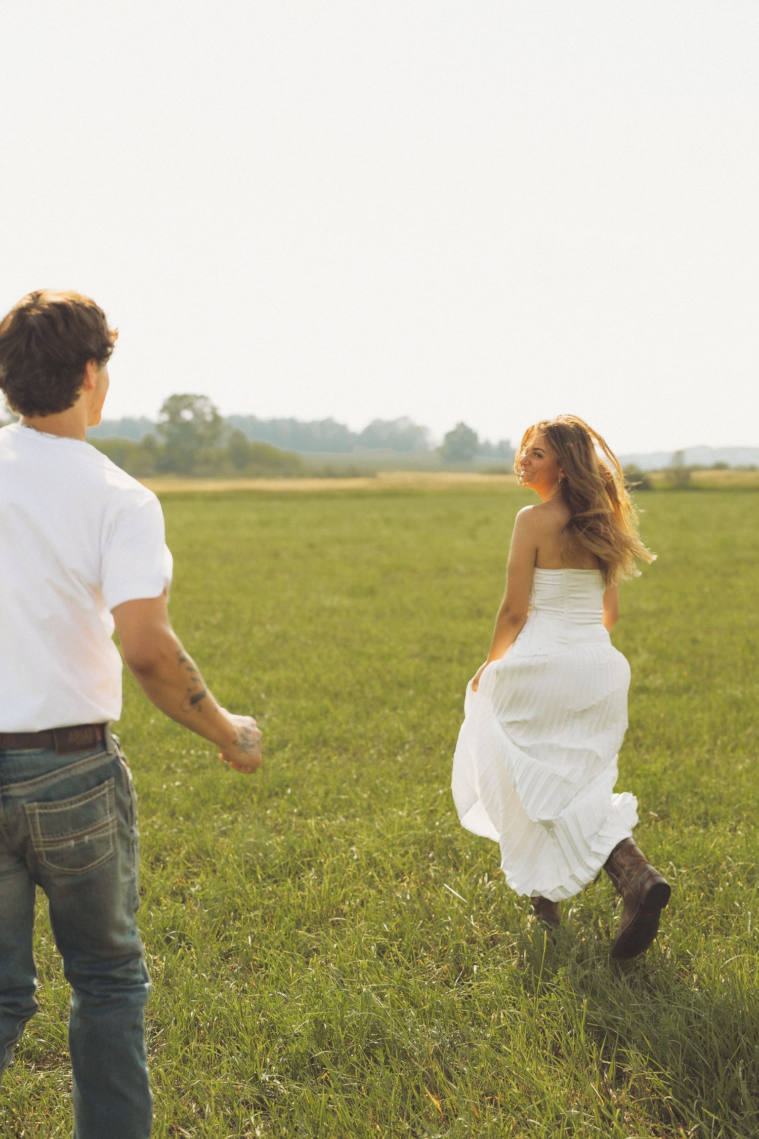 A couple runs through a green field at golden hour, woman in white dress and boots holding hands with man in white shirt.