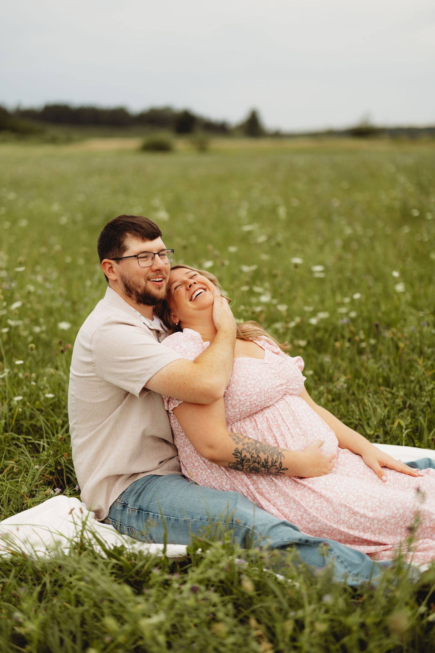 Couple sitting in a green field, pregnant woman in pink dress laughing joyfully while man embraces her.