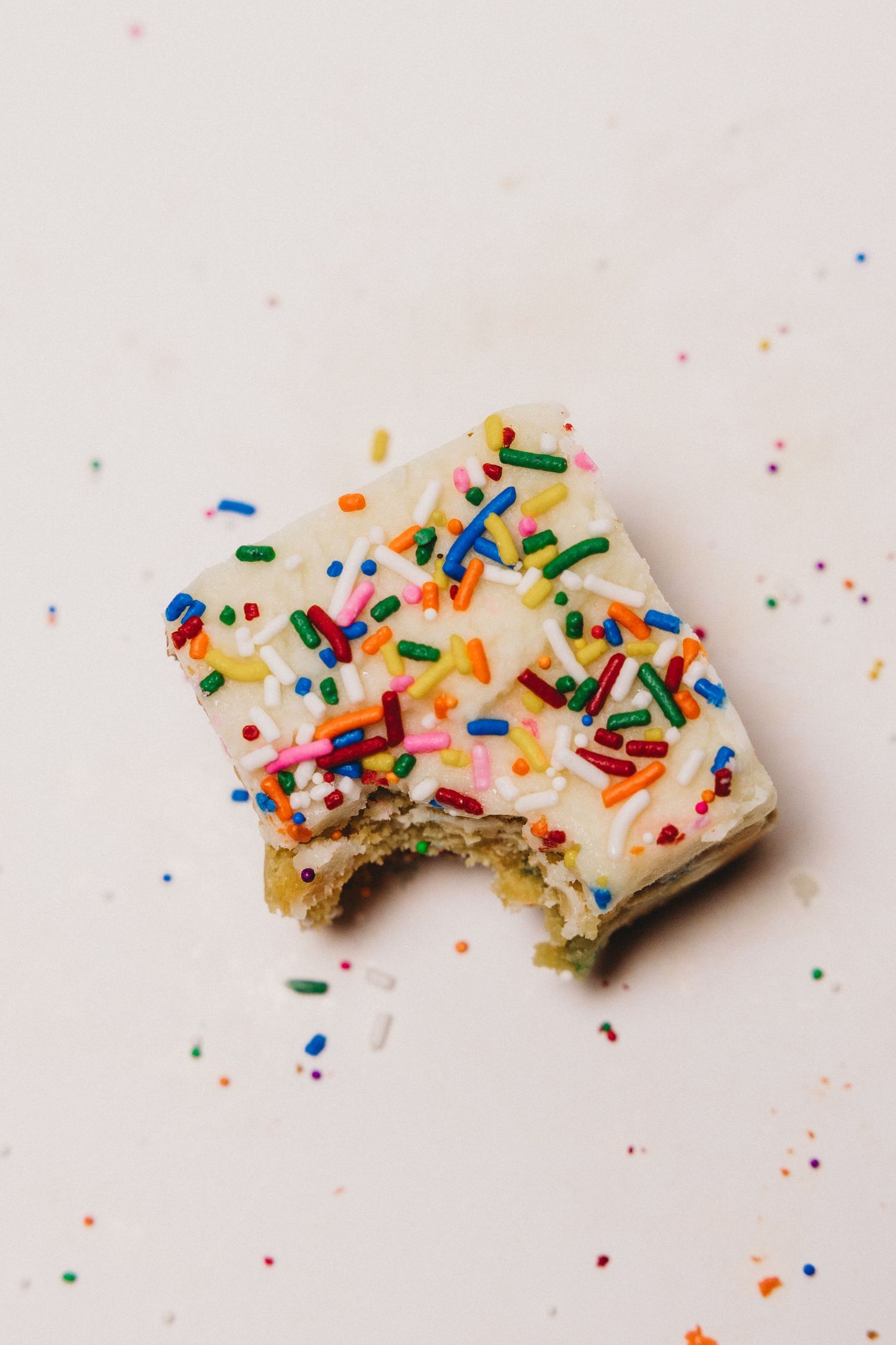 A frosted sugar cookie bar with colorful sprinkles, partially bitten, on a white surface.