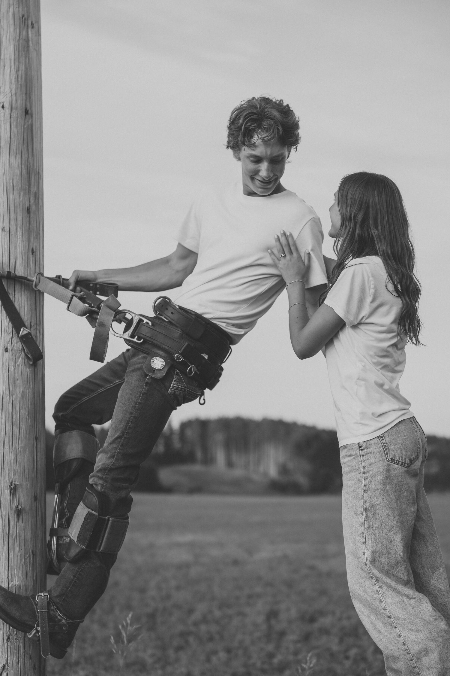 A lineman climbs a utility pole with climbing spurs while a young woman in jeans looks up at him smiling.