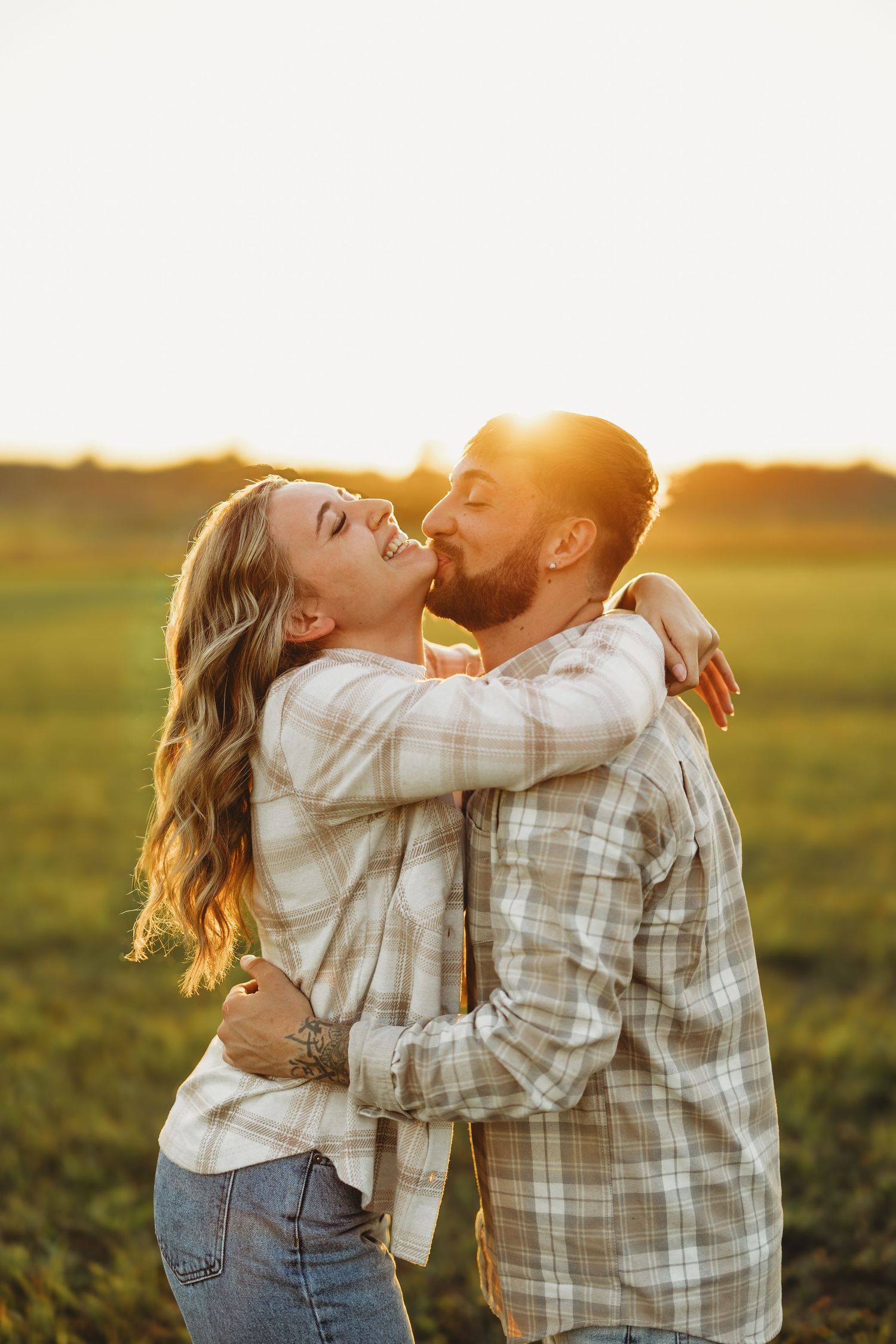 Couple embracing and kissing in a sunlit field at golden hour, both wearing matching plaid shirts.
