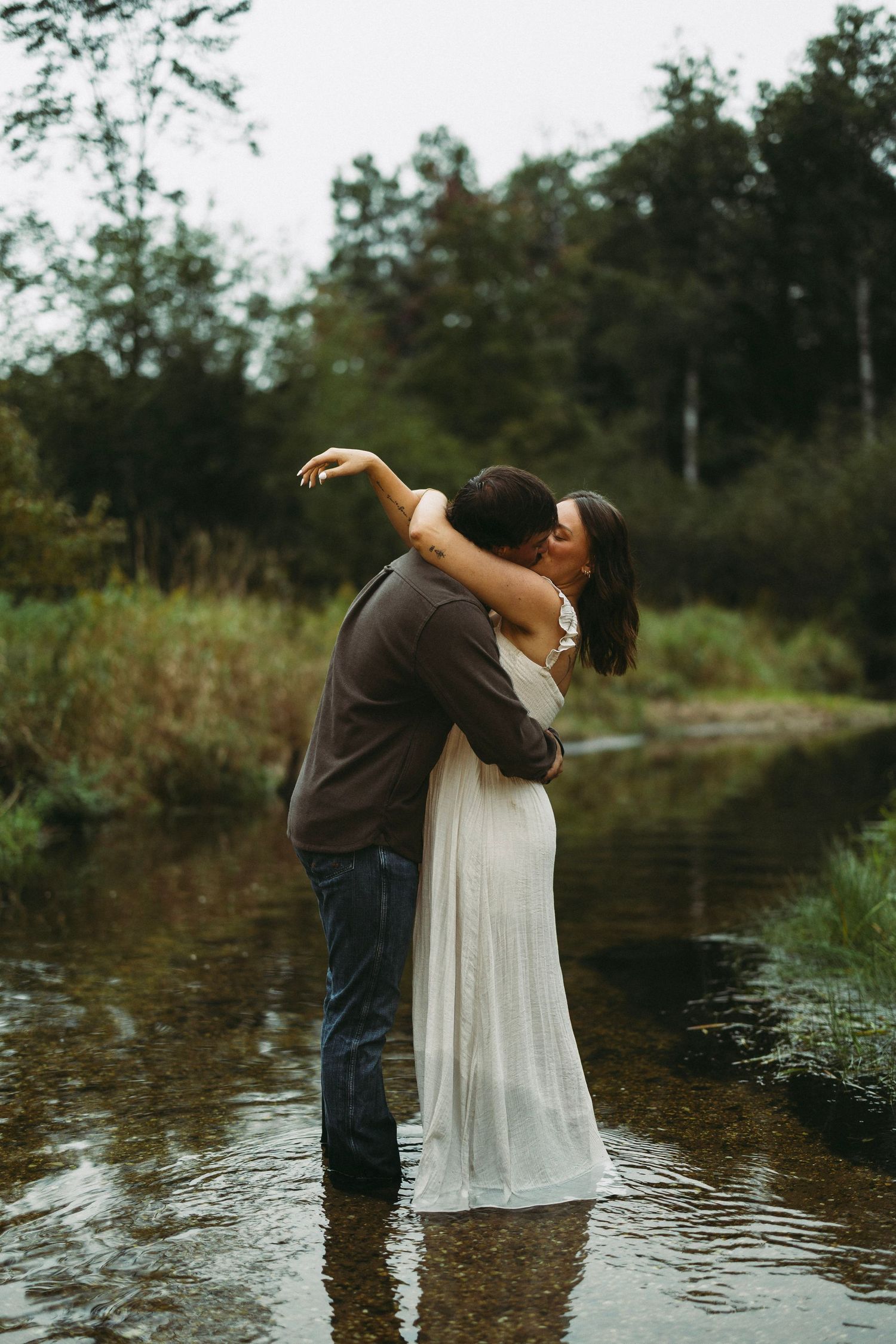 Couple in a long white dress and dark shirt kiss while standing in a shallow forest stream surrounded by green trees.