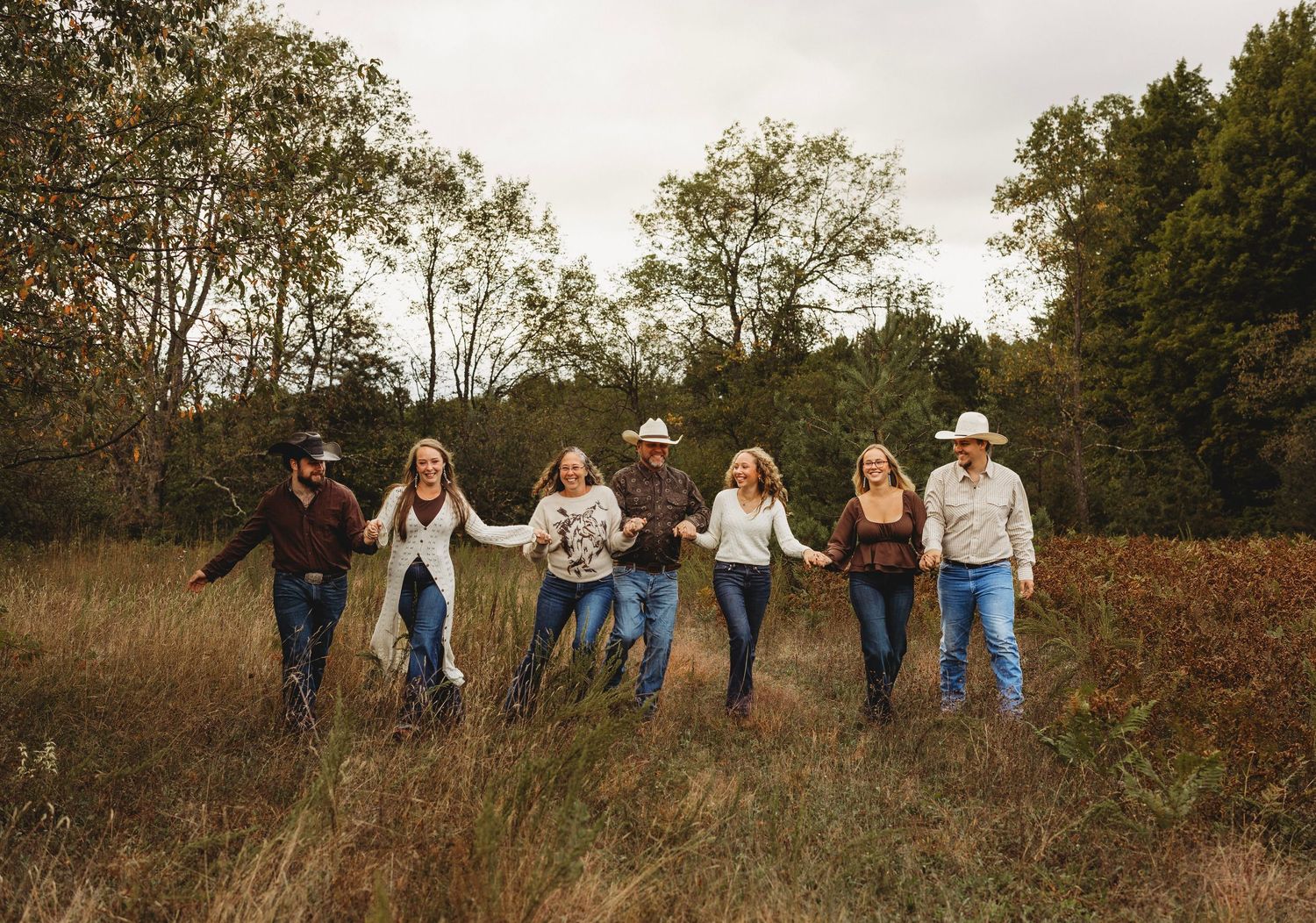 Family of eight in casual clothes and cowboy hats holding hands and walking through a golden autumn field with trees behind them.