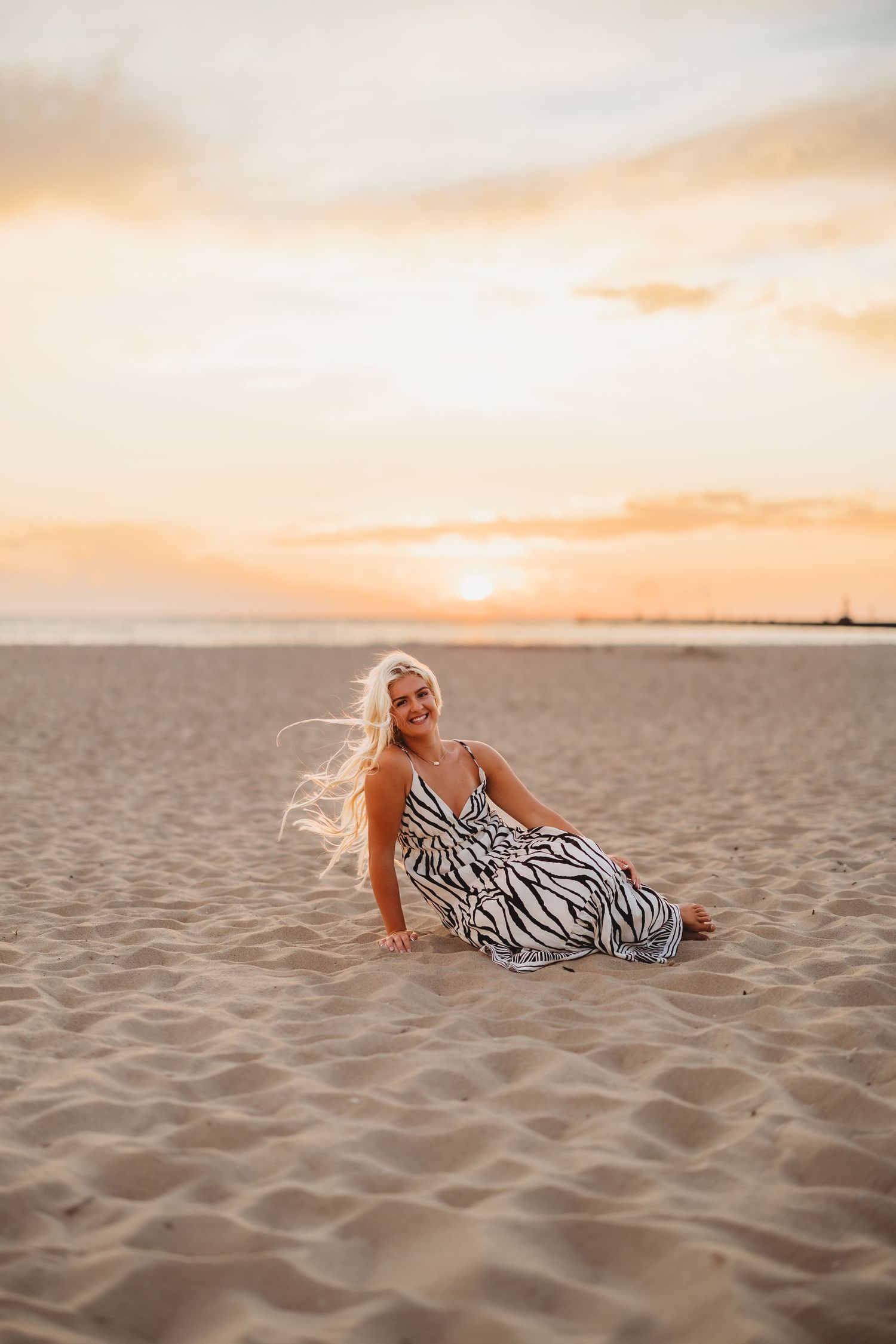 Blonde woman in a black and white zebra-print maxi dress sitting on a sandy beach at sunset with a glowing orange sky.