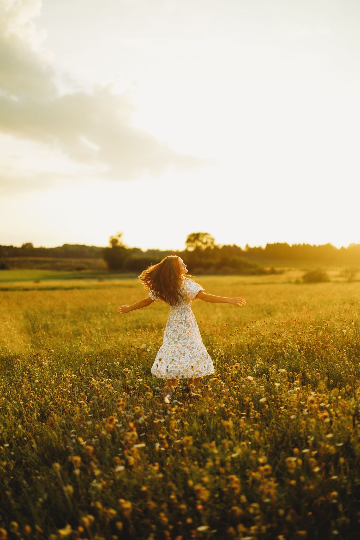 Girl in a white floral dress spinning with arms outstretched in a golden wildflower field at sunset.
