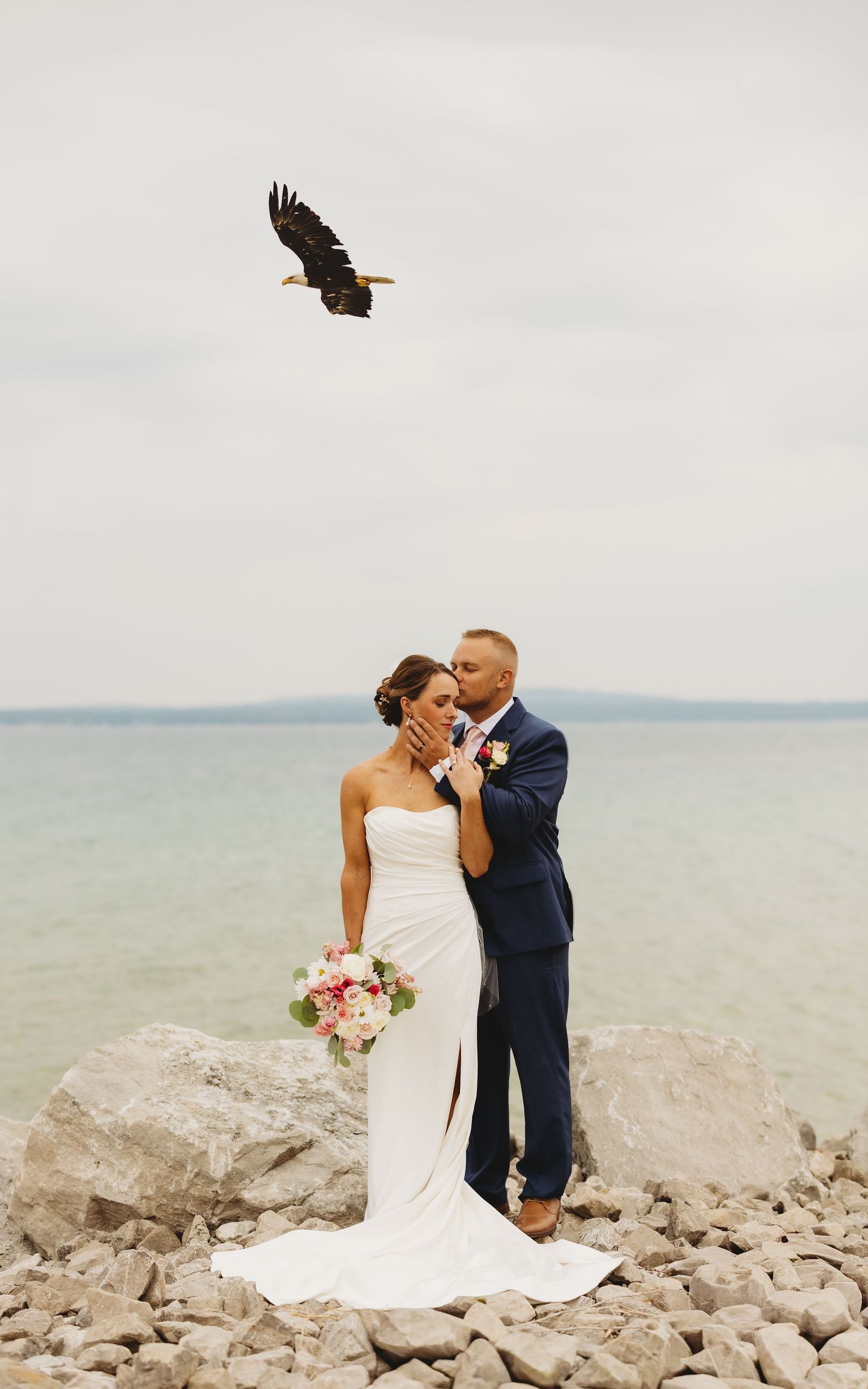 Bride and groom kissing on rocky lakeside shore in wedding attire with a bald eagle soaring overhead.