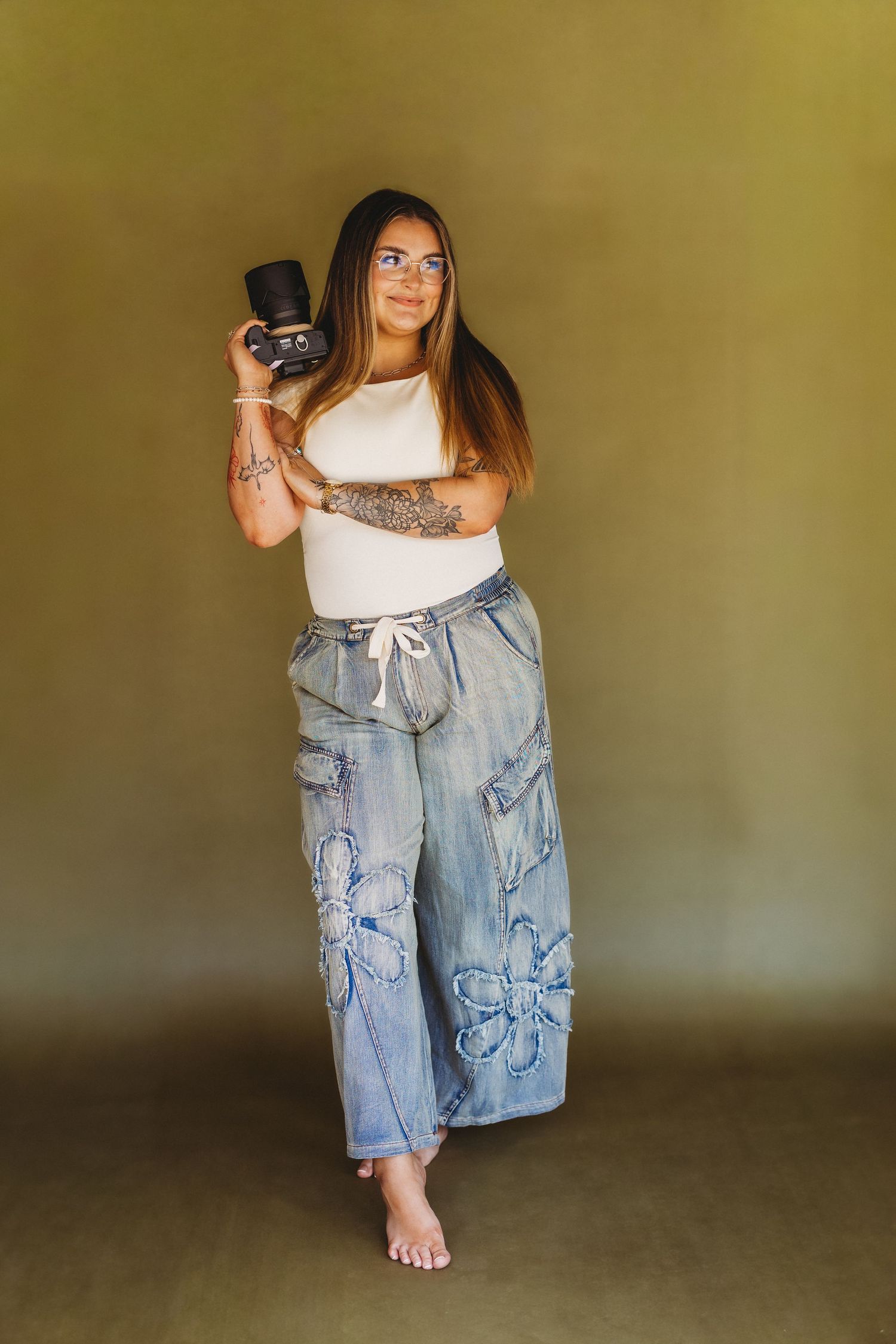 Tattooed woman in white top and wide-leg floral jeans holds camera, posing barefoot against olive background.