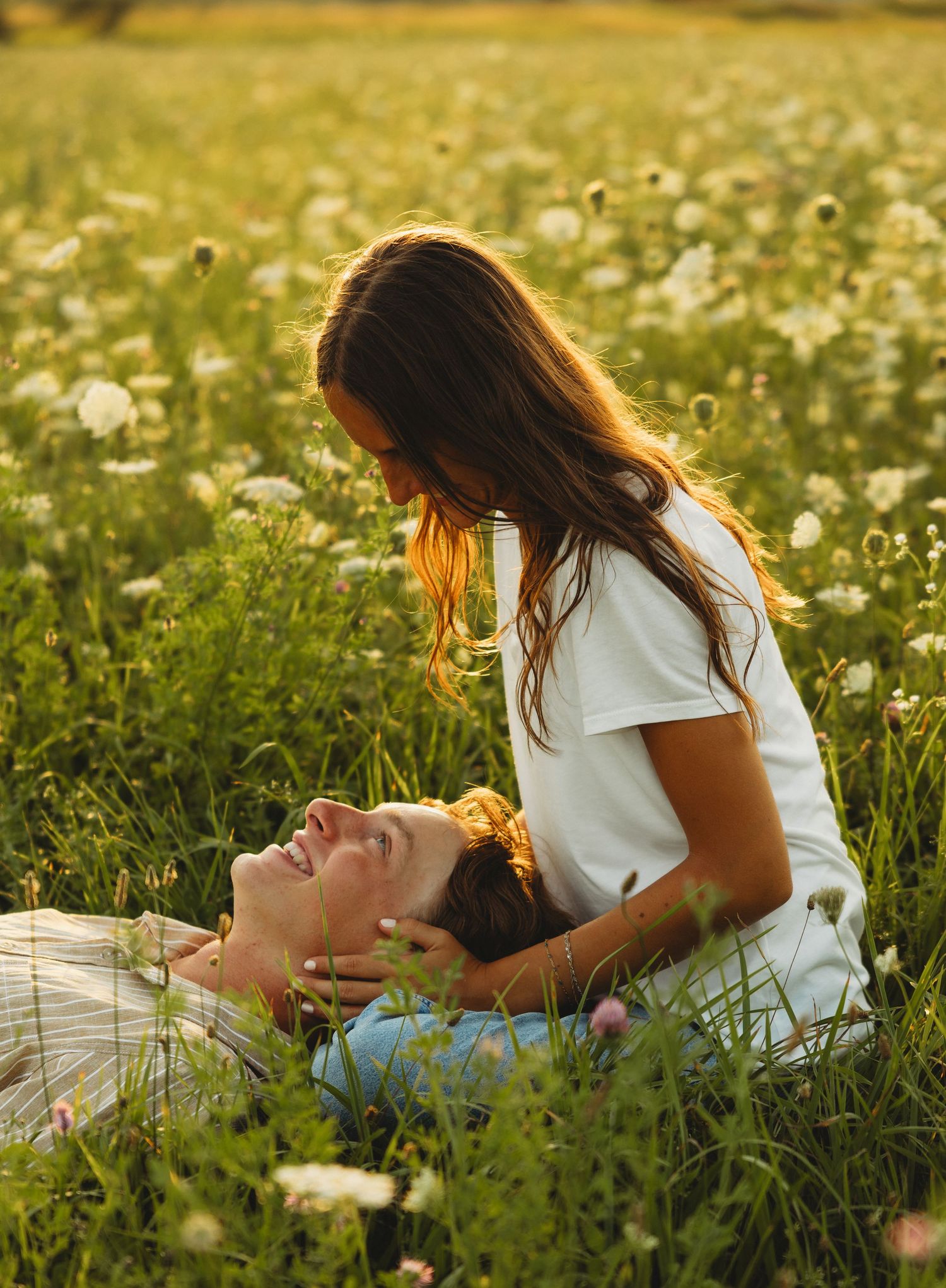 Romantic couple in a wildflower meadow, woman sitting beside man lying in grass during golden hour sunset.