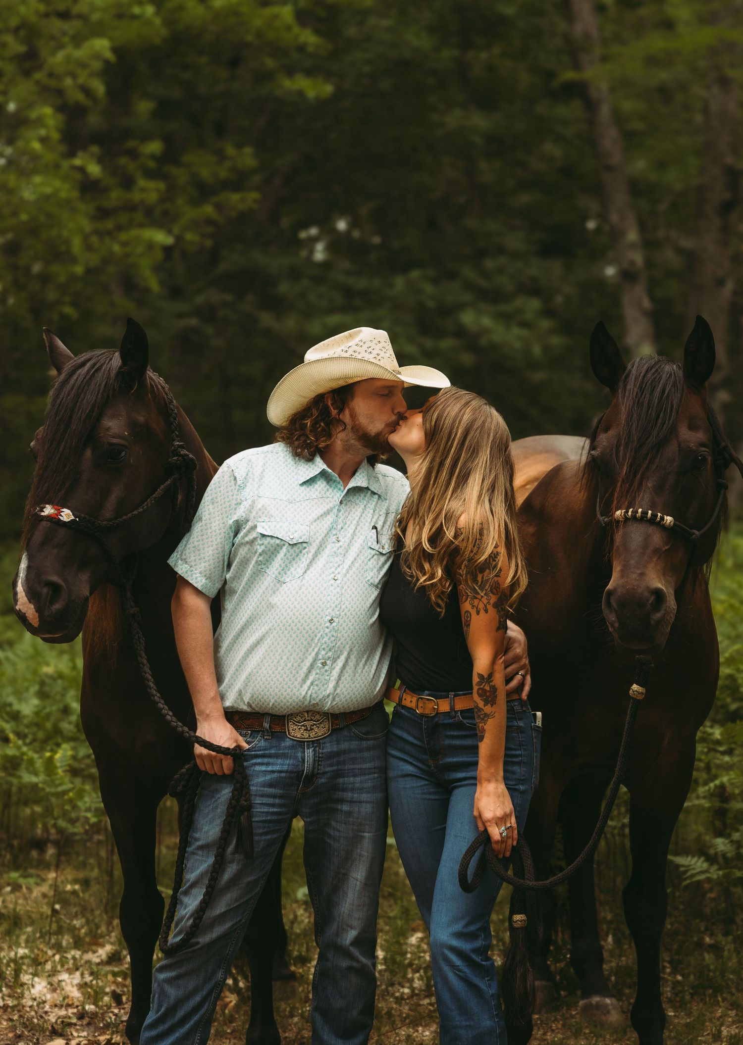 Couple kissing between two dark horses in a forest, man wearing cowboy hat and mint shirt, woman in black top and jeans.