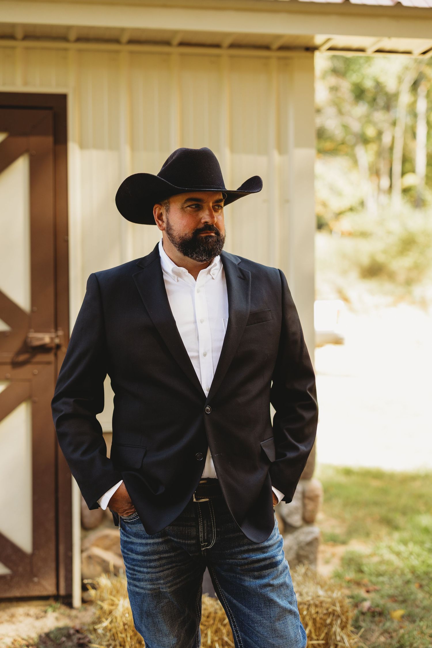 Bearded man in a black cowboy hat, dark blazer, white shirt and jeans stands beside a rustic barn door with hay at his feet.