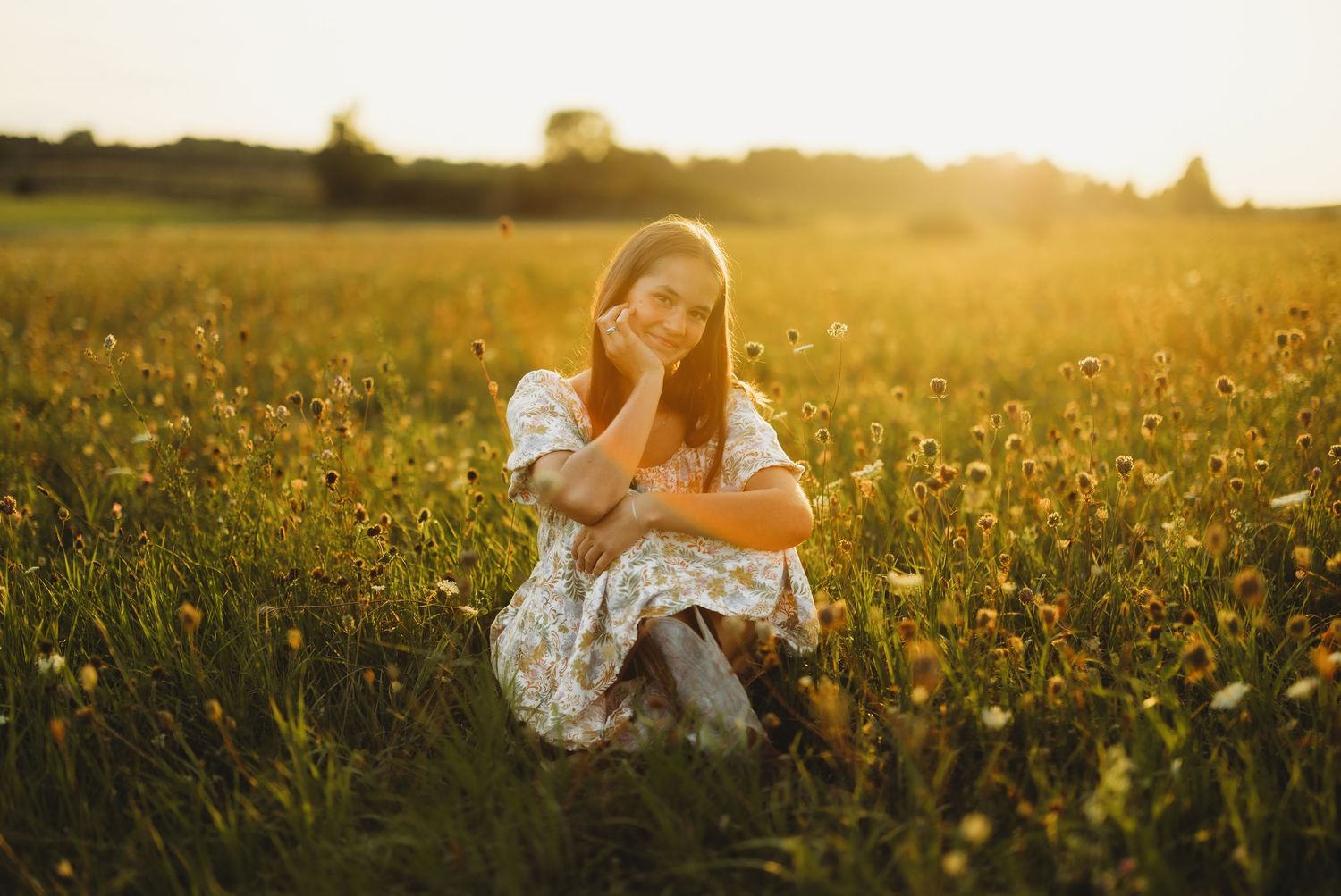 Young woman in a floral dress sits in a golden wildflower meadow at sunset, resting her chin on her hand and smiling softly.