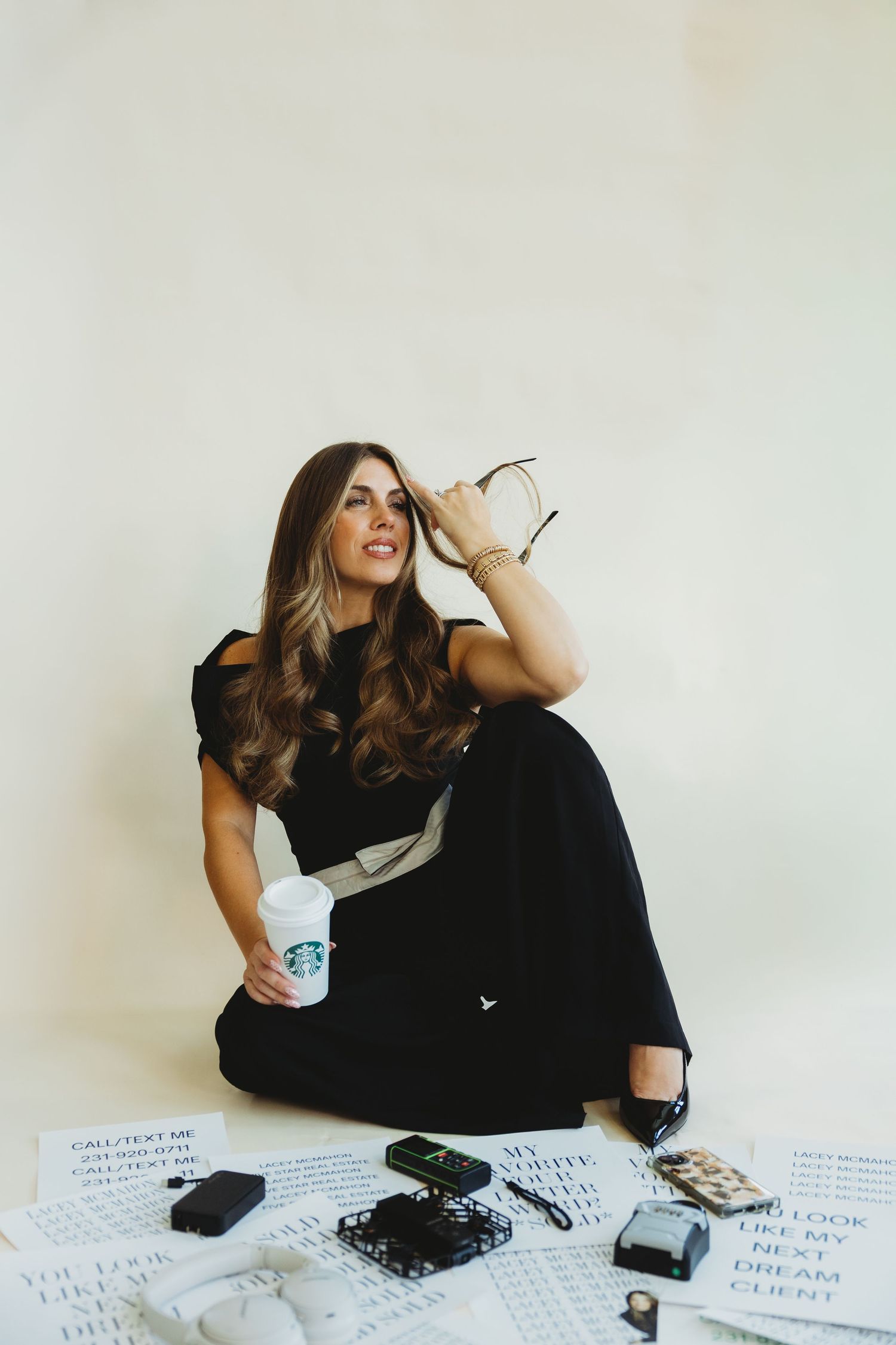 Woman in a black outfit sits on the floor holding glasses and a Starbucks cup, surrounded by papers, a phone, and desk accessories.