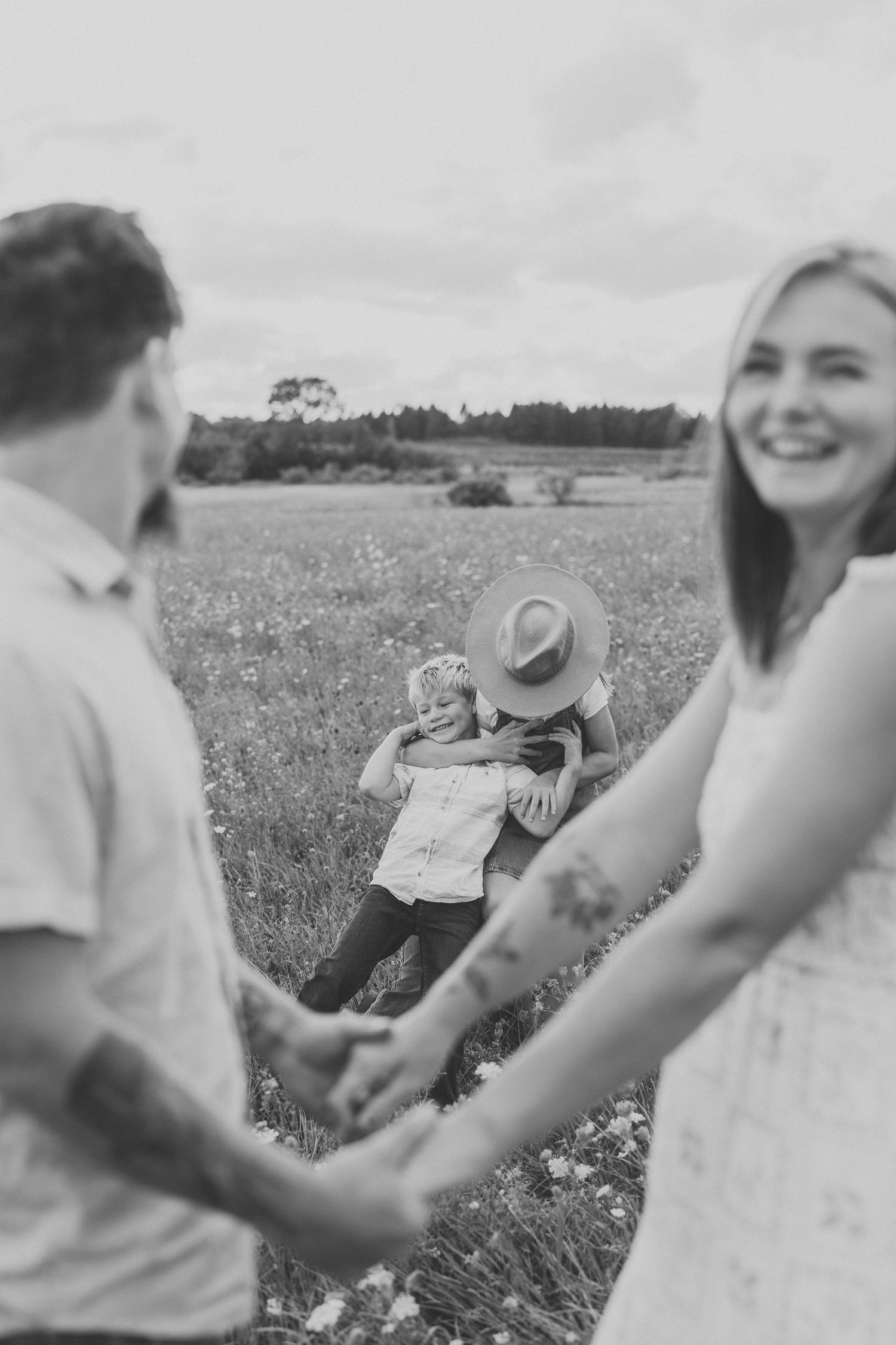 Black and white photo of a smiling couple holding hands in a wildflower field while a child in the background hugs a person in a sun hat.