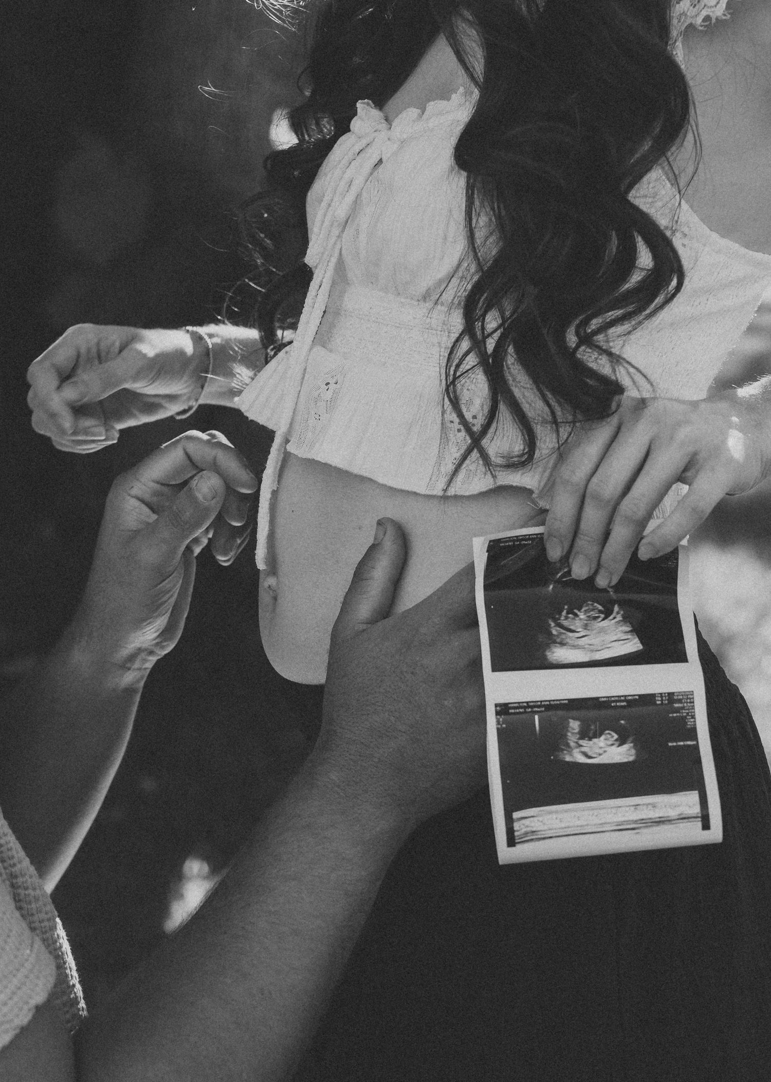 Black and white photo of pregnant woman holding ultrasound scan with partner touching her baby bump.