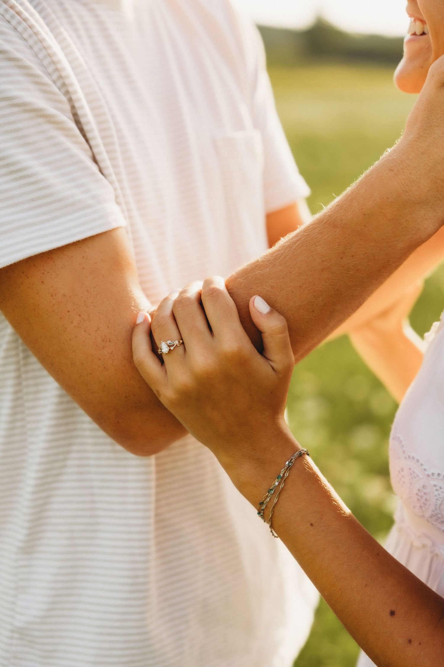 Close-up of couple holding hands outdoors, woman wearing engagement ring and bracelet in golden sunlight.