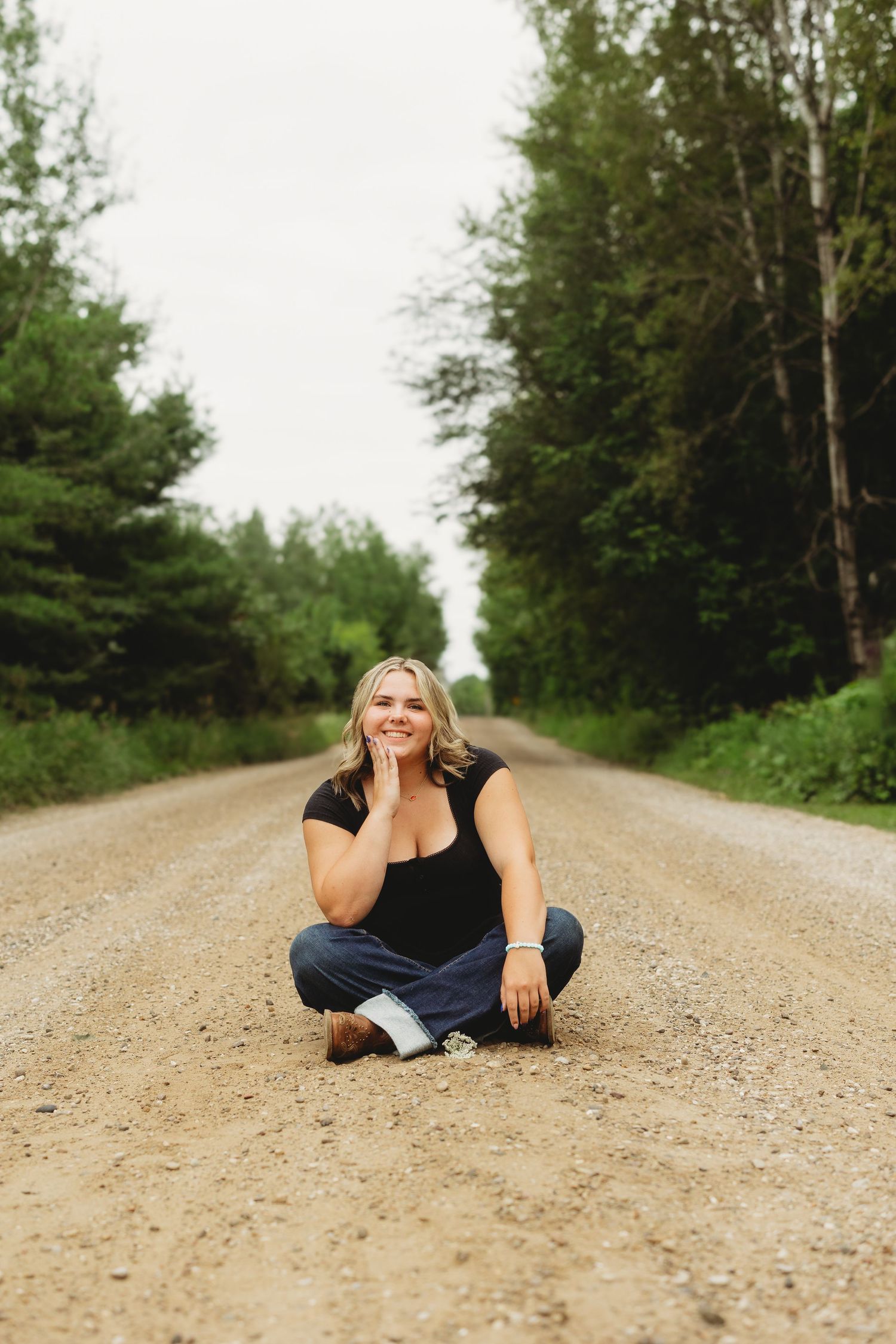 Smiling blonde woman sitting cross-legged on dirt road surrounded by lush green trees.