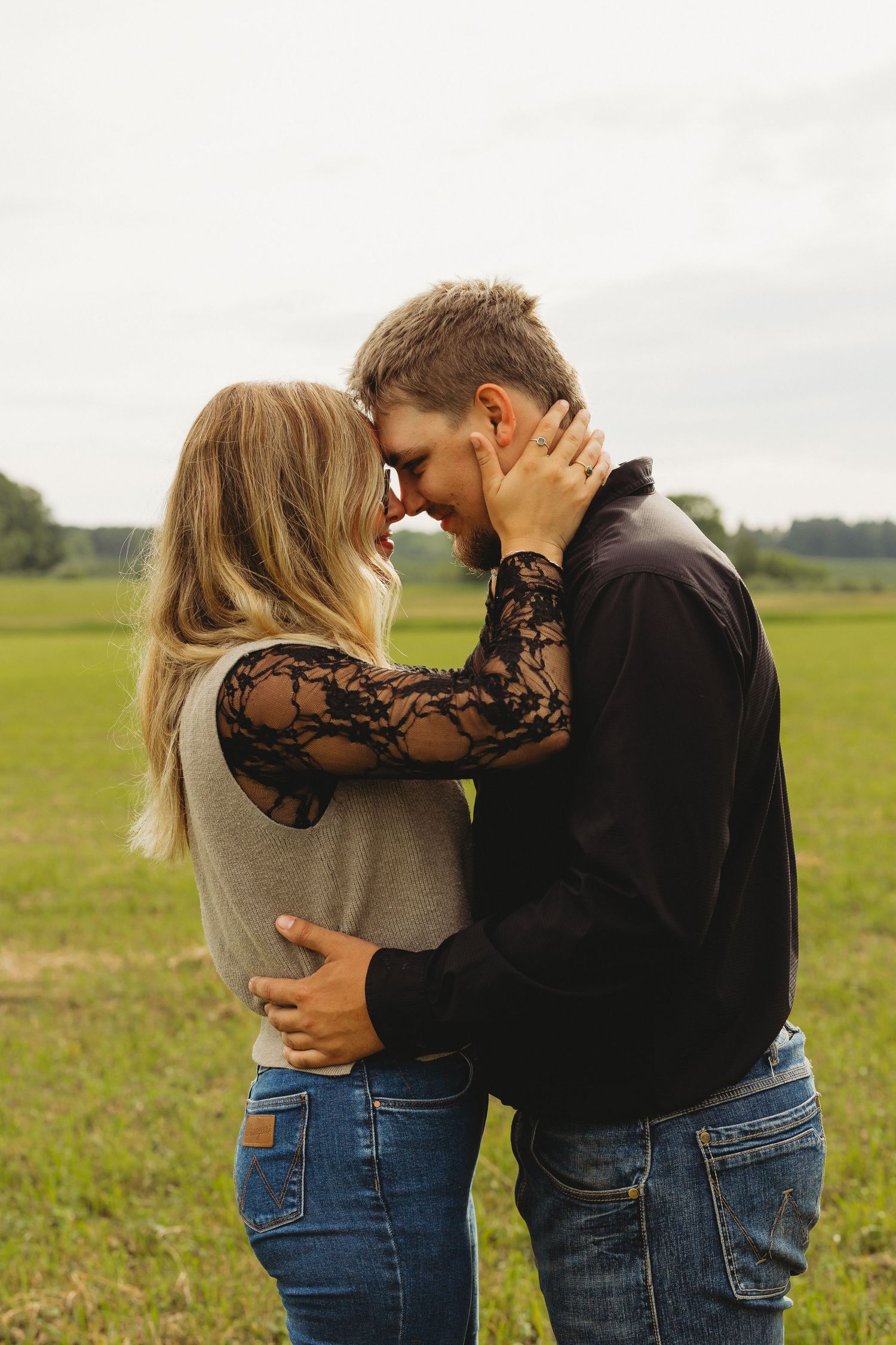 Romantic couple embracing forehead to forehead in a green field, woman in lace sleeve top, man in black shirt.