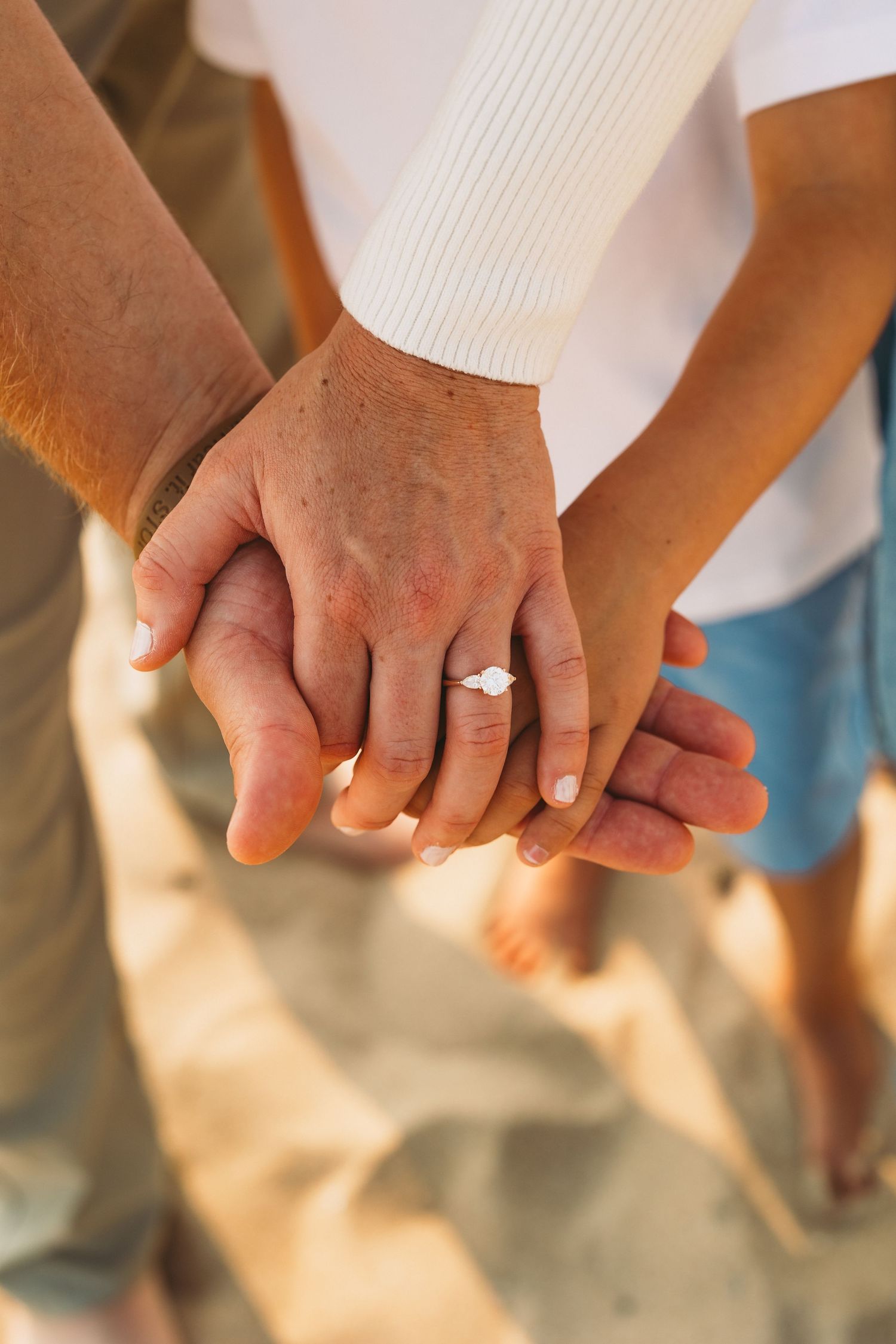 Close-up of hands stacked on sandy beach, woman wearing diamond engagement ring, symbolizing love and commitment.