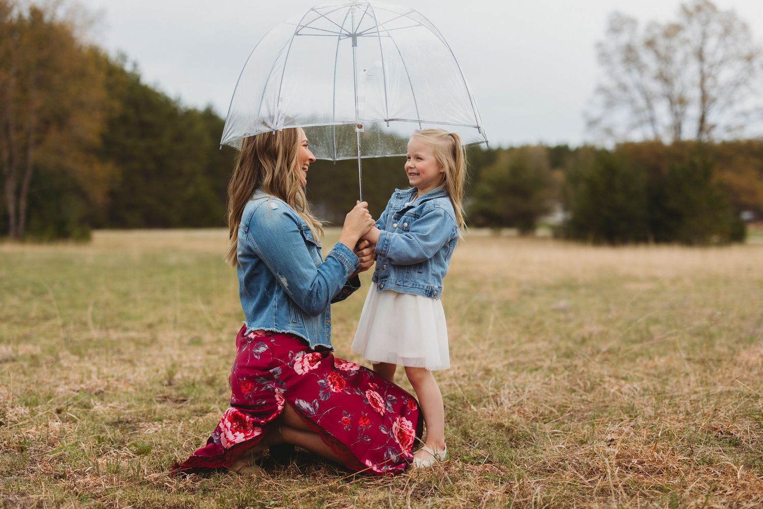 Mother and daughter share a clear umbrella in a field, both wearing denim jackets, sharing a sweet moment.