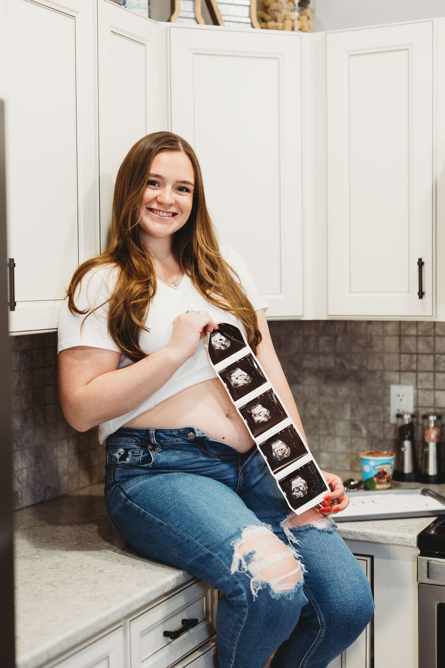 Smiling pregnant woman sitting on kitchen counter holding ultrasound strip, wearing white top and ripped jeans.