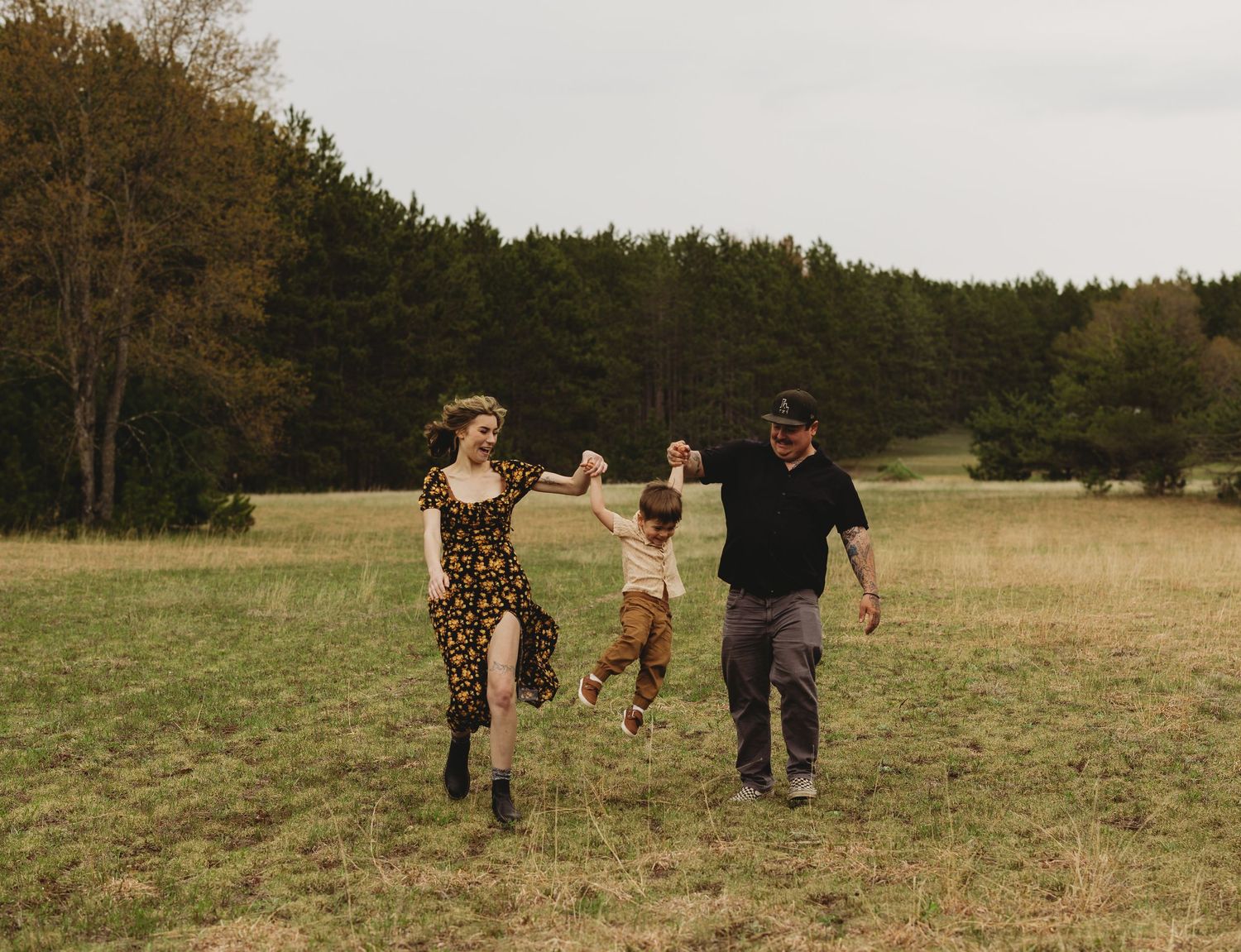 Family of three playing in a green meadow, parents swinging child between them with forest backdrop.