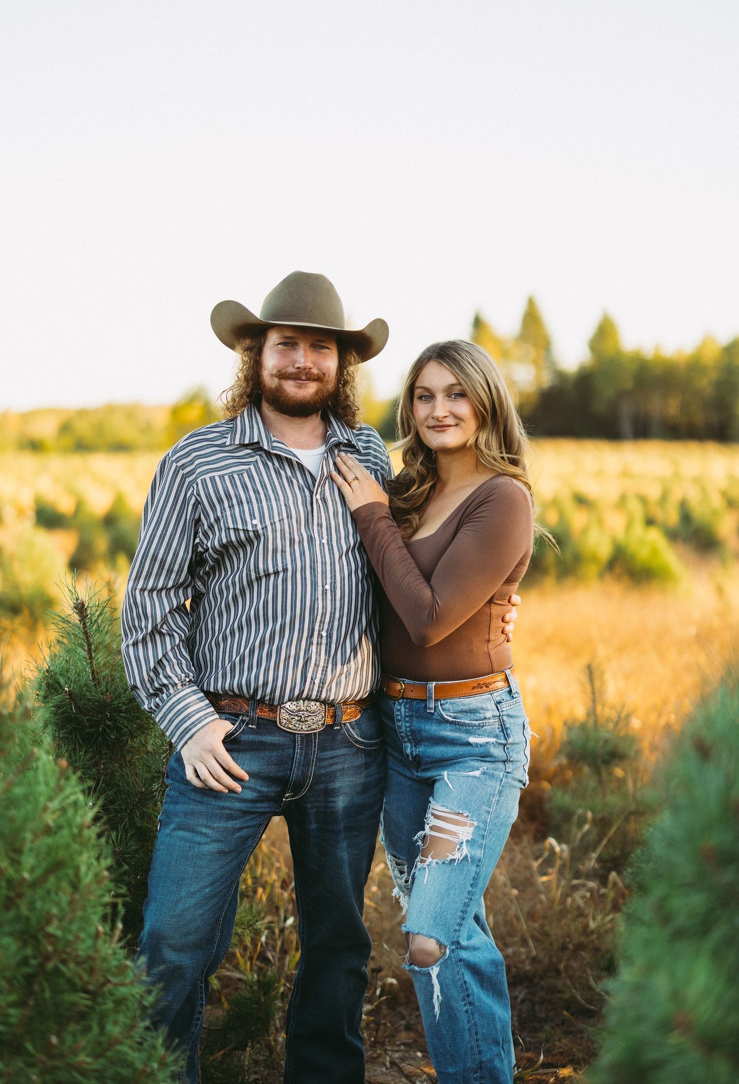 A couple in western attire poses together in a golden field at sunset, wearing denim and casual clothing.