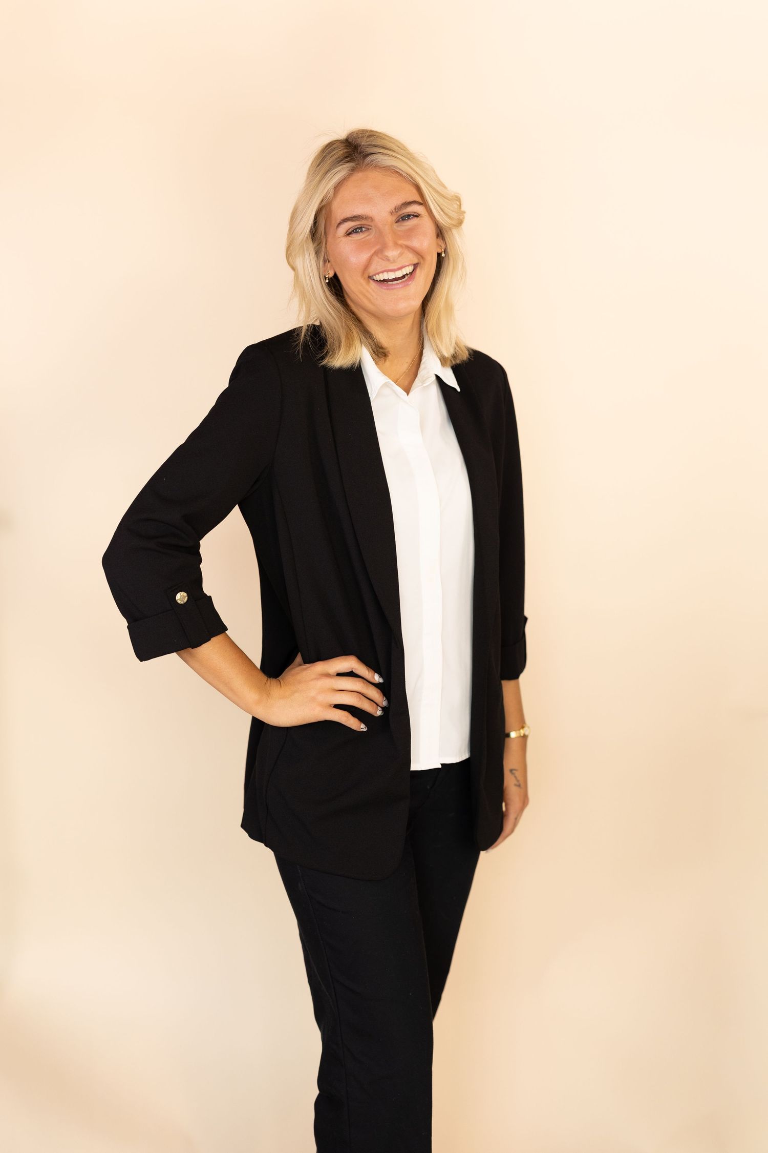 Smiling blonde woman in black blazer and white shirt poses confidently against a neutral beige background.