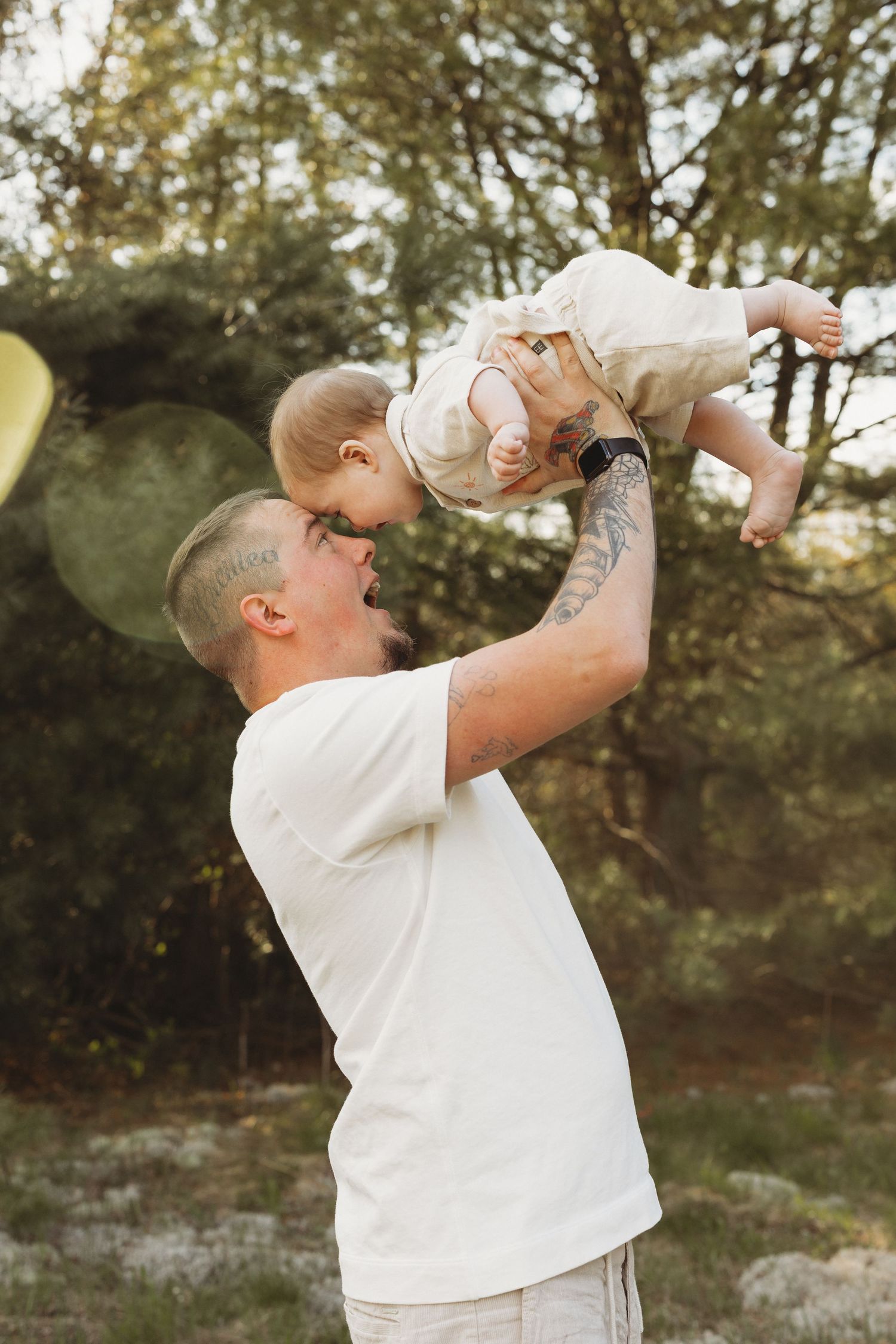 Tattooed father in white t-shirt lifting smiling baby overhead in outdoor garden setting.