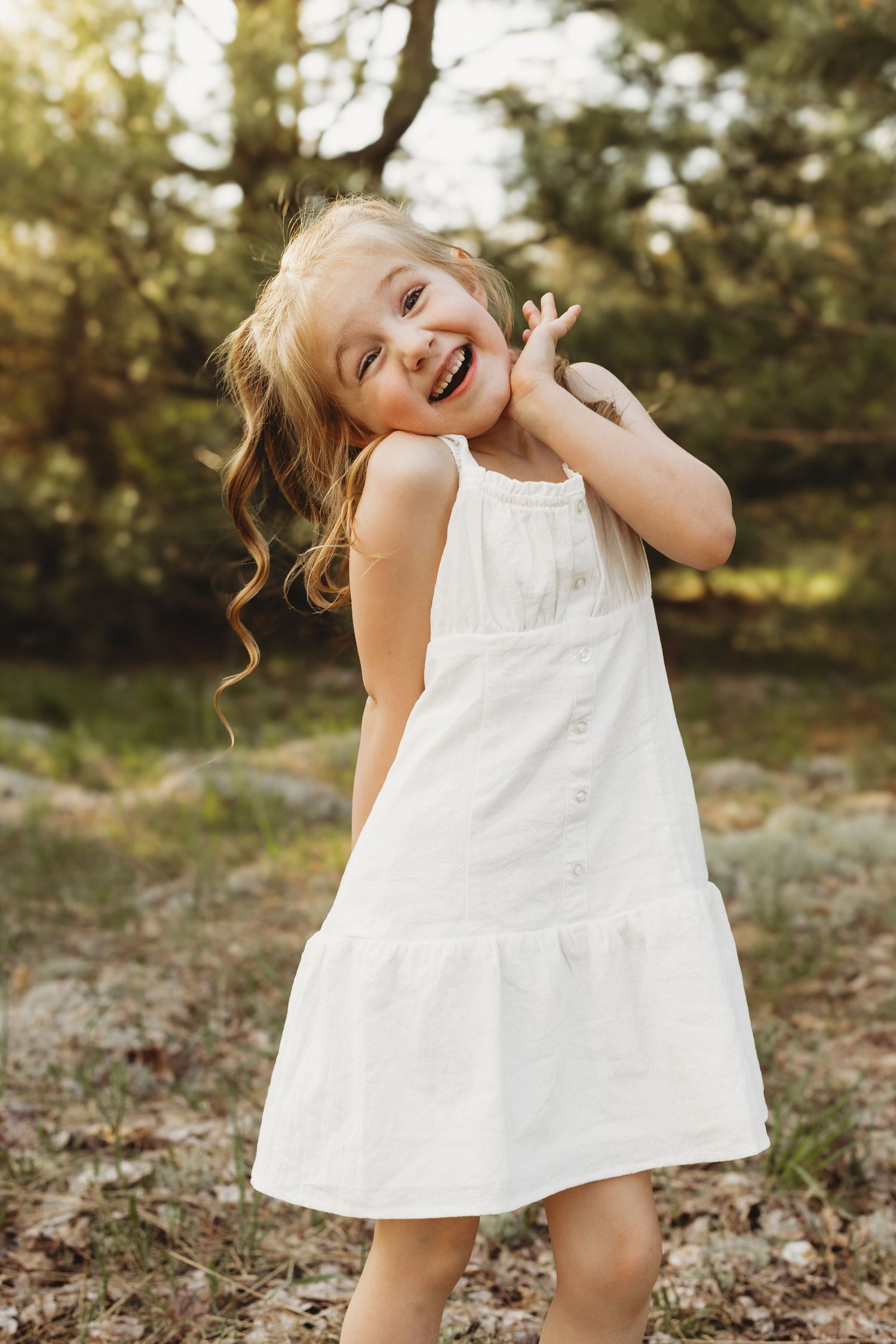 A joyful young girl with curly blonde hair laughs outdoors wearing a white sundress in a sunlit forest setting.