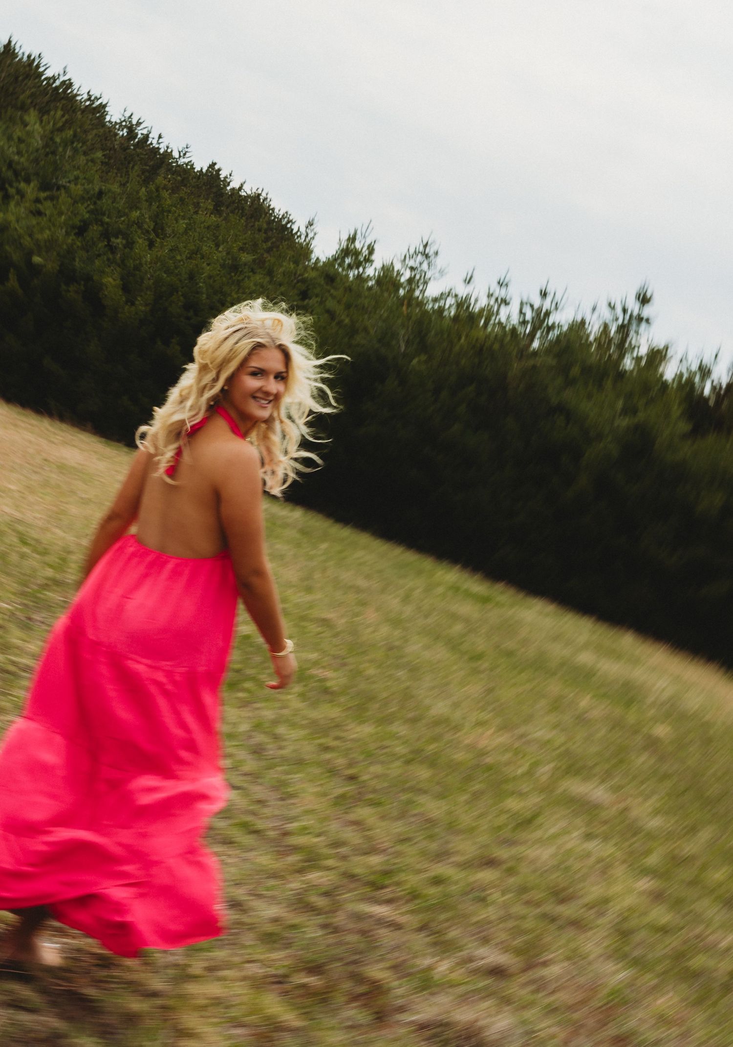 Blonde woman in pink halter dress running through green field with trees in background.