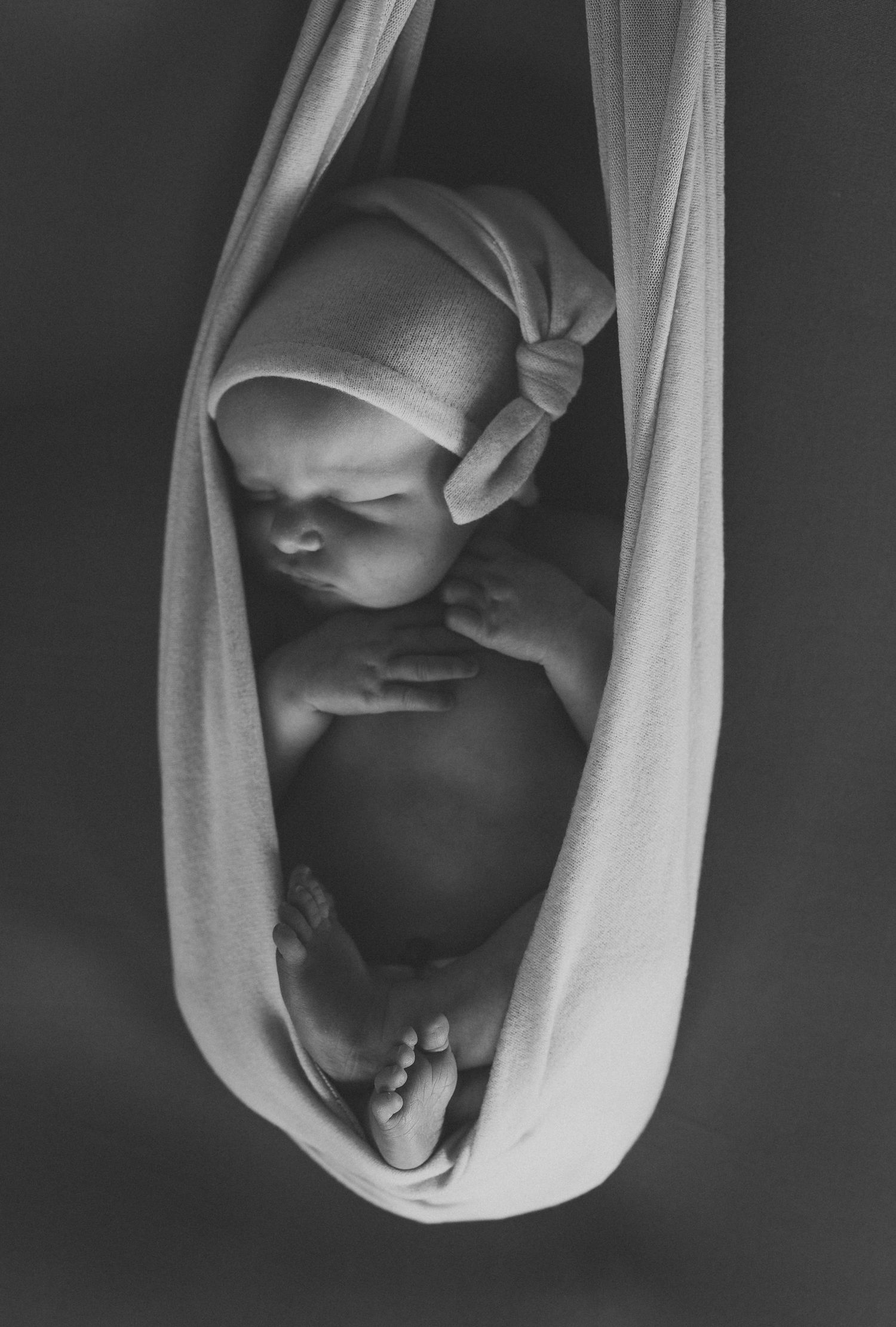 Black and white photo of a sleeping newborn baby cradled in a soft white fabric sling, wearing a tiny knit hat.